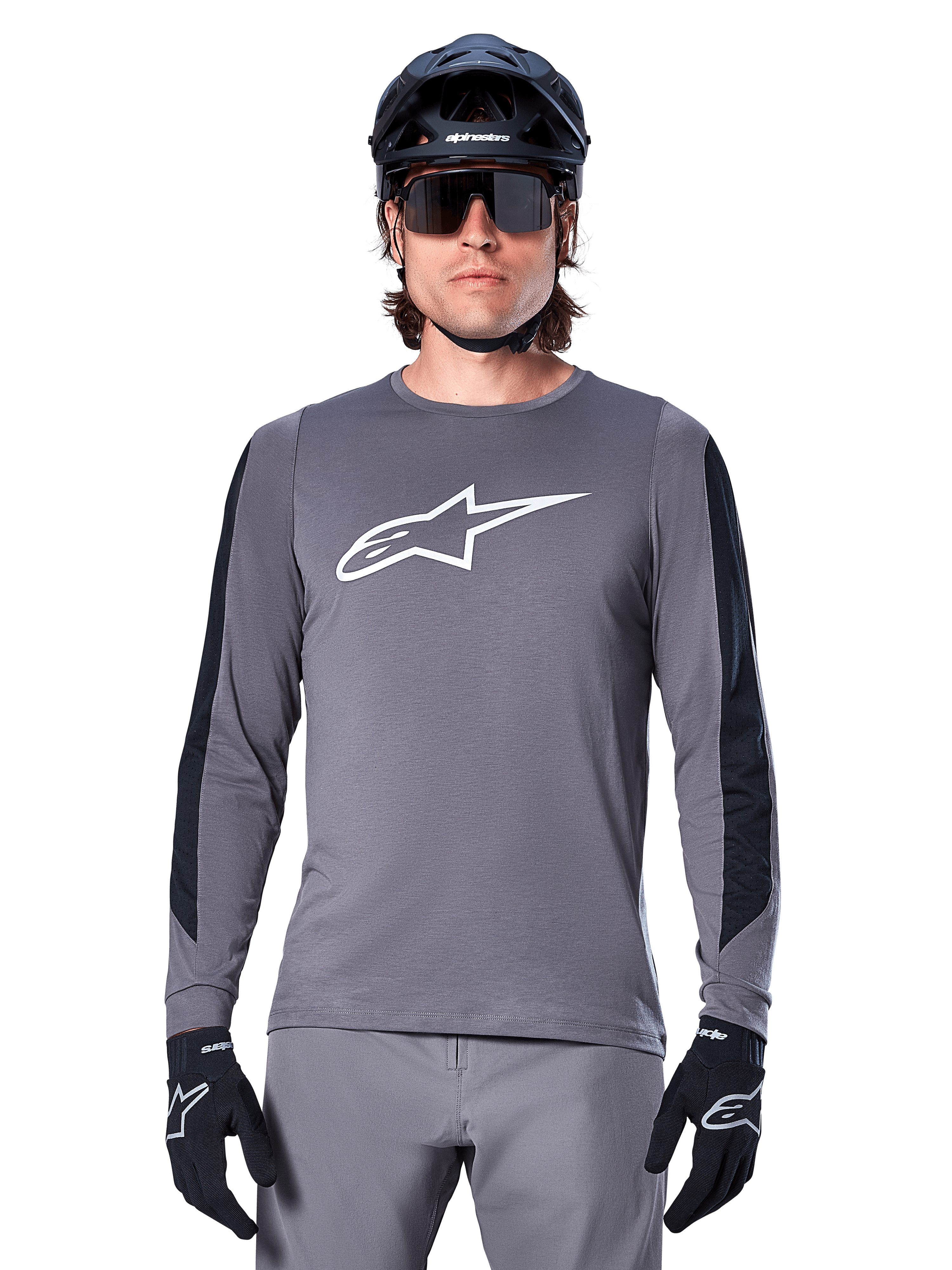 Alpinestars A-Dura Dri Astar Jersey met lange mouwen, donkergrijs, voorzien van een groot wit Astar-logo op de borst en zwarte mesh-panelen op de mouwen, gedragen door een rijder met een zwarte mountainbikehelm en sportzonnebril, MTB-fietskleding