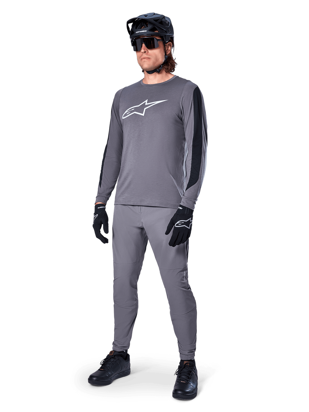 Alpinestars A-Dura Dri Astar Jersey, MTB-shirt met lange mouwen, donkergrijs met wit logo op de borst, zwarte mesh-panelen op de mouwen, gedragen met bijpassende grijze broek, zwarte helm en handschoenen, performance mountainbike-kleding
