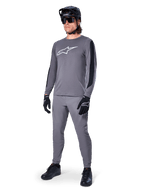 Alpinestars A-Dura Dri Astar Jersey, MTB-shirt met lange mouwen, donkergrijs met wit logo op de borst, zwarte mesh-panelen op de mouwen, gedragen met bijpassende grijze broek, zwarte helm en handschoenen, performance mountainbike-kleding