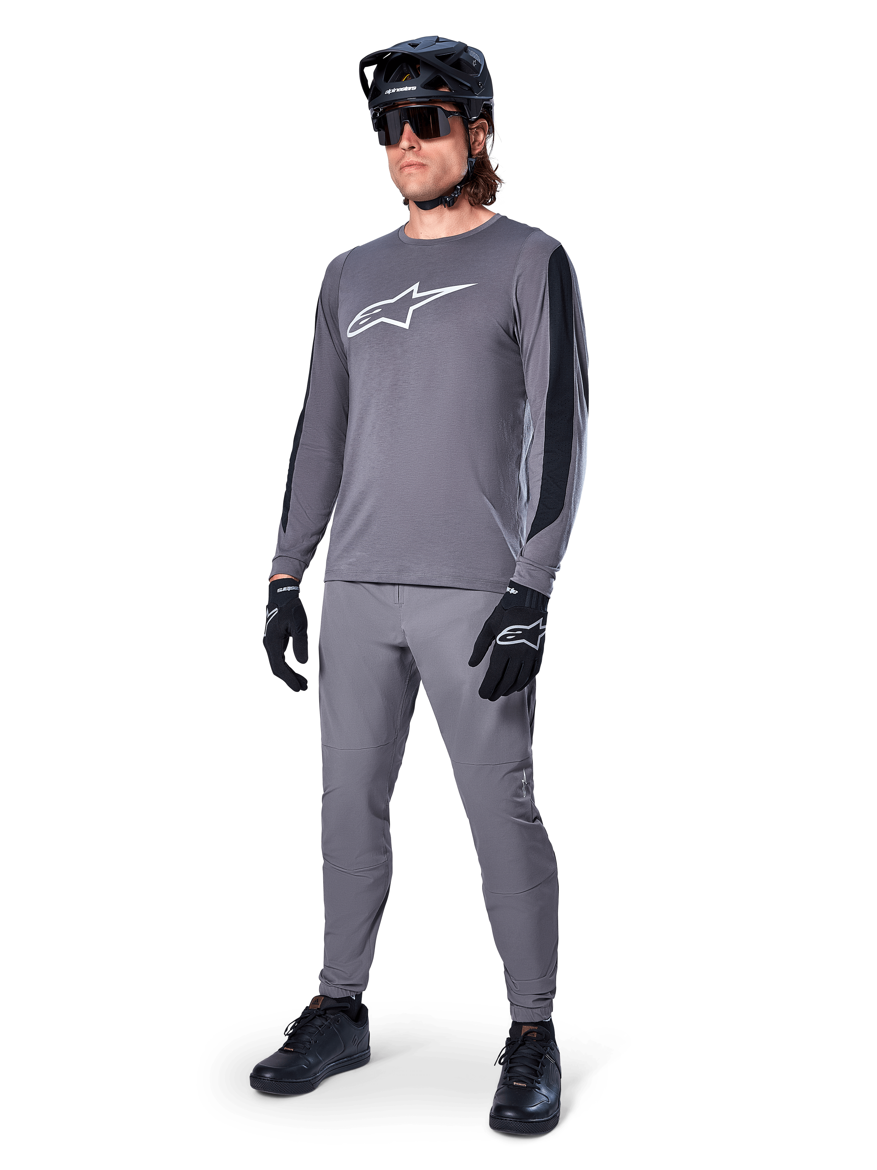 Alpinestars A-Dura Dri Astar Jersey, MTB-shirt met lange mouwen, donkergrijs met wit logo op de borst, zwarte mesh-panelen op de mouwen, gedragen met bijpassende grijze broek, zwarte helm en handschoenen, performance mountainbike-kleding