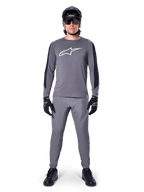 Alpinestars A-Dura Dri Astar Jersey, MTB-shirt met lange mouwen in donkergrijs, voorzien van een wit logo op de borst en zwarte mouwpanelen, gedragen met een bijpassende grijze broek, zwarte helm, zonnebril en handschoenen voor een complete off-road fietsoutfit