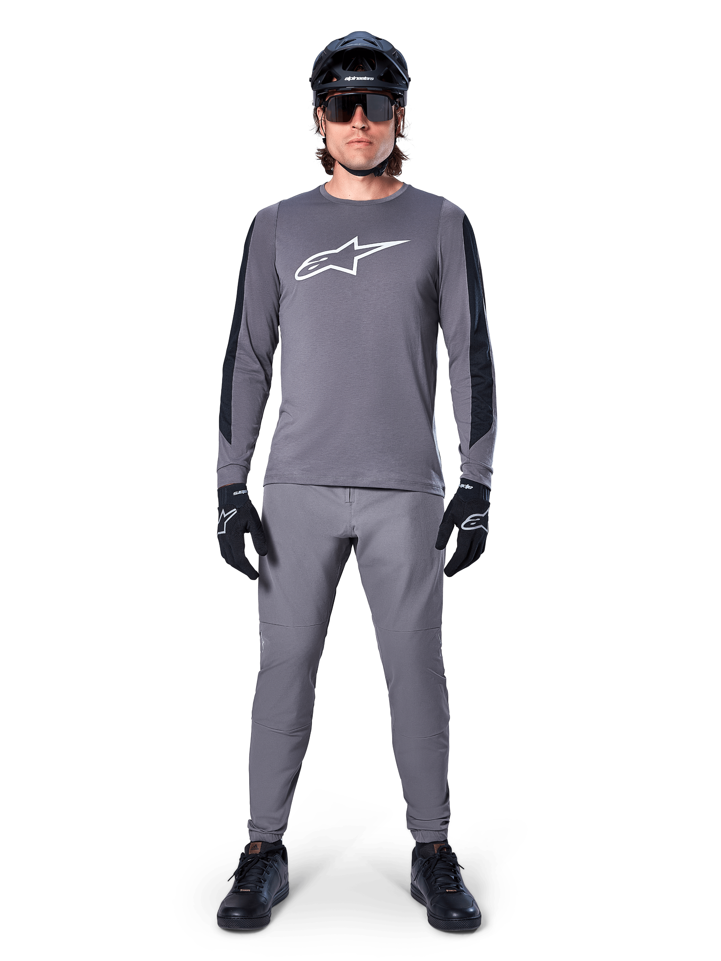 Alpinestars A-Dura Dri Astar Jersey, MTB-shirt met lange mouwen in donkergrijs, voorzien van een wit logo op de borst en zwarte mouwpanelen, gedragen met een bijpassende grijze broek, zwarte helm, zonnebril en handschoenen voor een complete off-road fietsoutfit