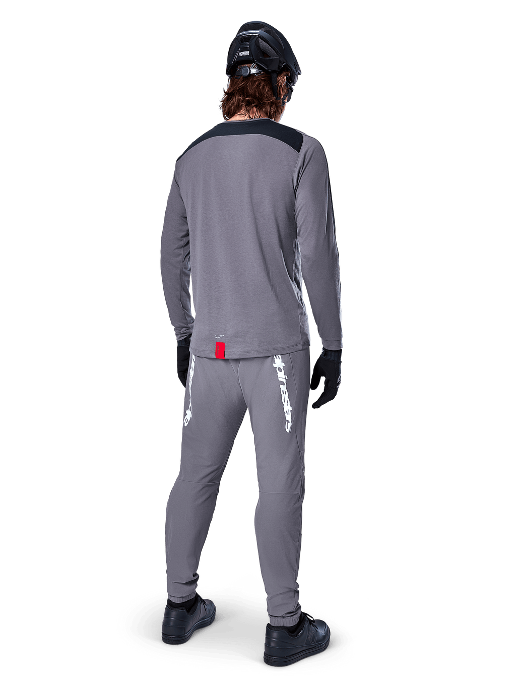 Alpinestars A-Dura Dri Astar Jersey met lange mouwen, donkergrijs, achteraanzicht van een rijder met bijpassende broek en zwarte helm, voorzien van zwarte schouderpanelen en een klein rood label aan de zoom, technische off-road fietskleding