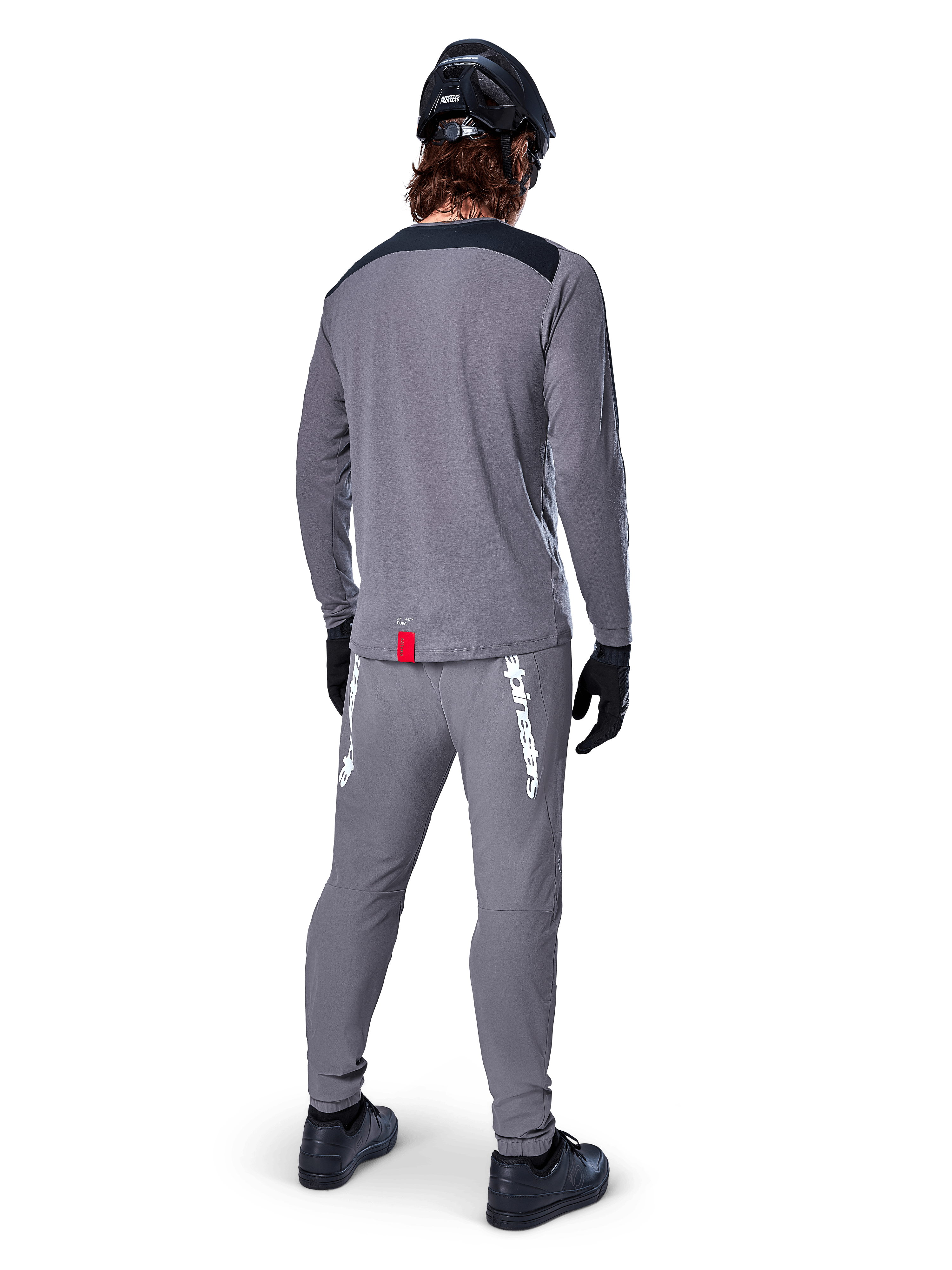 Alpinestars A-Dura Dri Astar Jersey met lange mouwen, donkergrijs, achteraanzicht van een rijder met bijpassende broek en zwarte helm, voorzien van zwarte schouderpanelen en een klein rood label aan de zoom, technische off-road fietskleding