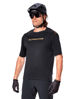 Alpinestars A-Aria Polartec Switch Trikot Kurzarm, schwarzes Mountainbike-Trikot mit goldenem Alpinestars-Branding, getragen mit schwarzem Helm, Sonnenbrille und Handschuhen, technische MTB-Bekleidung