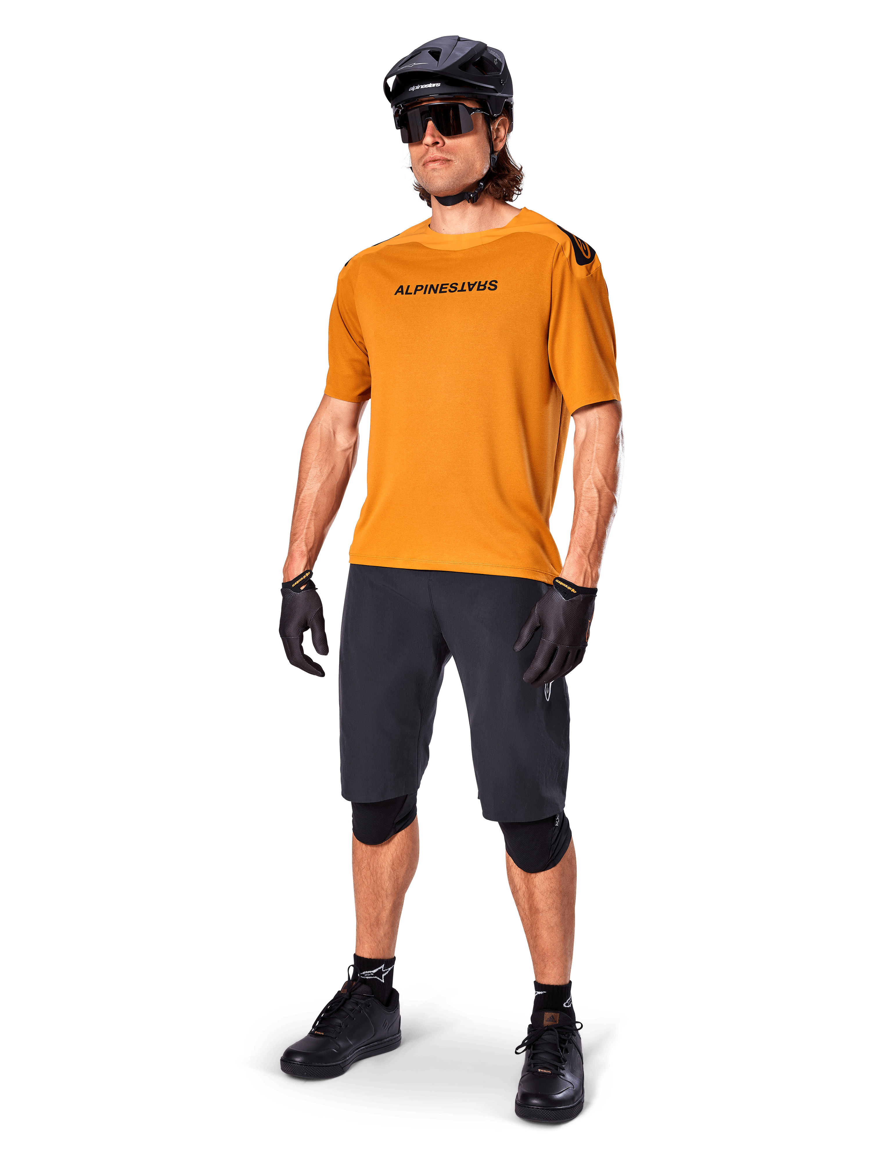 Male rider wearing the Alpinestars A-Aria Polartec Switch Jersey in Dark Gold, short-sleeve MTB jersey, paired with black cycling shorts, knee guards, black helmet, and gloves, professional mountain bike outfit for trail riding