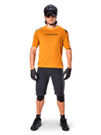 Male cyclist wearing the Alpinestars A-Aria Polartec Switch Short Sleeve Jersey in Dark Gold, paired with black MTB shorts, helmet, sunglasses, and gloves, professional mountain bike trail riding gear