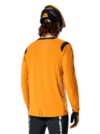 Alpinestars A-Dura Dri Wool Jersey met lange mouwen in Dark Gold, achteraanzicht van een rijder met mountainbikehelm en handschoenen, voorzien van zwarte mesh-panelen op de schouders en een verlengde achterzijde, technische fietskleding voor trailrijden