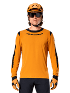 Mannelijk model draagt Alpinestars A-Dura Dri Wool Jersey met lange mouwen in Dark Gold, gecombineerd met een bijpassende gouden MTB-helm en handschoenen, zwarte mesh panelen op schouders en mouwen, zwart Alpinestars-woordmerk op de borst, donkere zonnebril en broek, technische mountainbikekleding