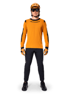 Mountainbiker met Alpinestars A-Dura Dri Wool jersey met lange mouwen, donkergoud met zwarte accenten op de kraag en mouwen, gecombineerd met een zwarte broek, bijpassende gouden helm, handschoenen en zwarte zonnebril, professionele mountainbike-uitrusting