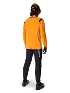 Mountainbiker van achteren gezien, dragend het Alpinestars A-Dura Dri Wool Jersey met lange mouwen in Dark Gold, zwarte technische broek met witte logo's en een bijpassende zwart-oranje helm, prestatiegerichte mountainbikekleding.