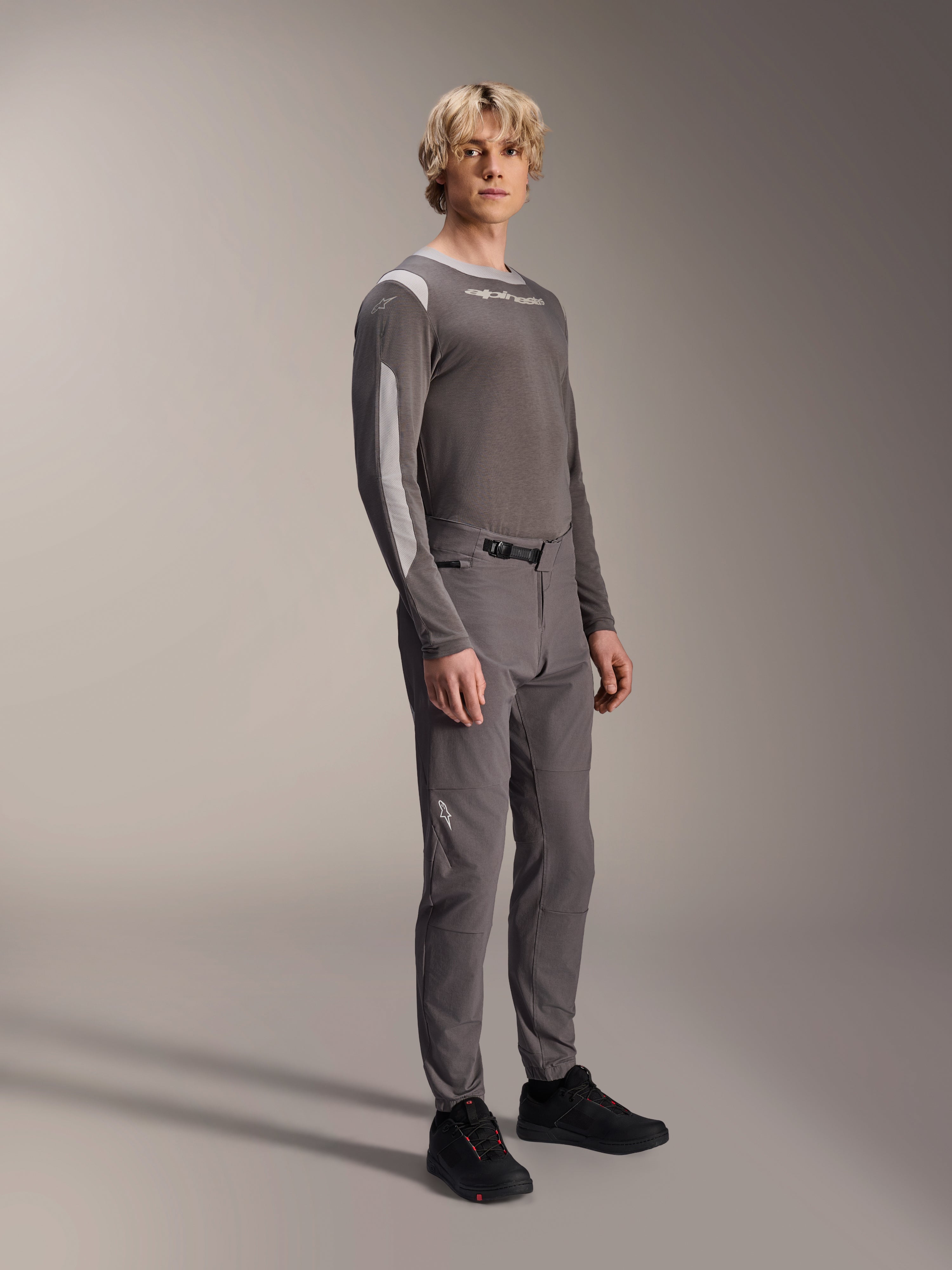 Alpinestars A-Dura Elite Pants, mountainbikebroek, donkergrijs, gedragen met een bijpassende jersey met lange mouwen en zwarte schoenen, met een ratelsluiting aan de voorzijde en duurzame Cordura-stof voor trail- en enduro-ritten.