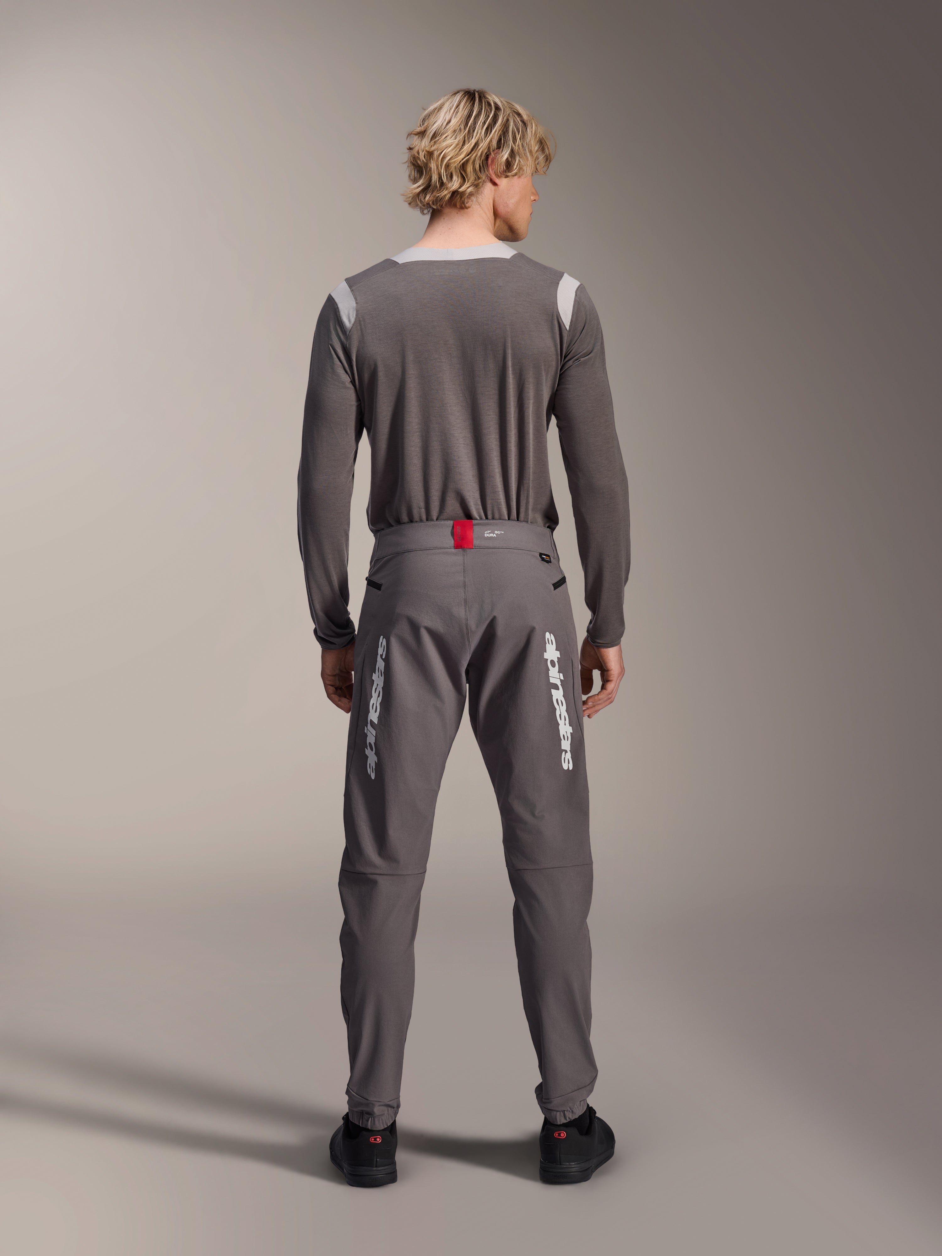 Alpinestars A-Dura Elite Pants in donkergrijs, model van achteren gezien met bijpassende grijze jersey met lange mouwen, witte Alpinestars-logo's op de pijpen, rood accent op de tailleband, zwarte schoenen, professionele MTB-uitrusting