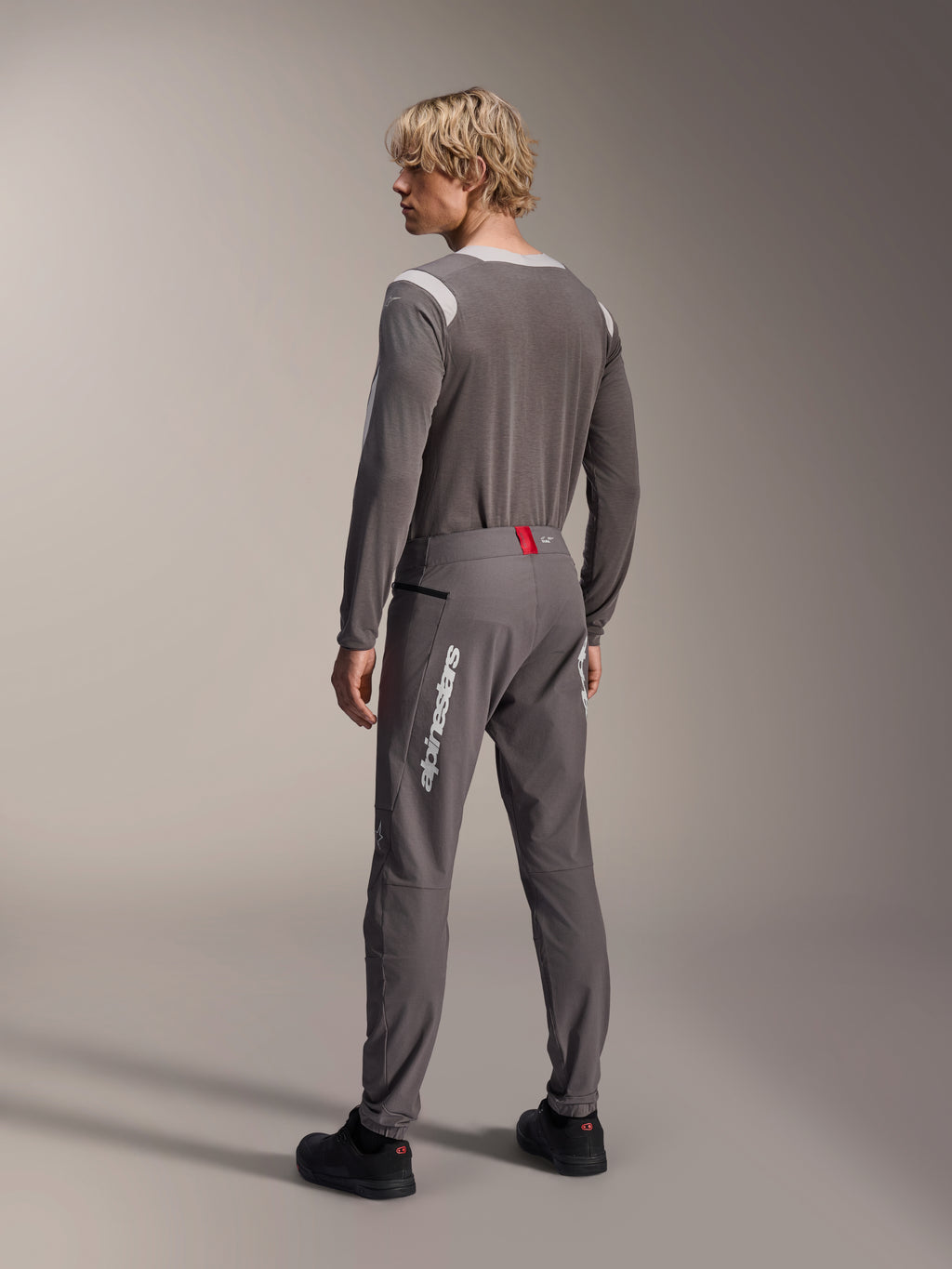 Model van achteren gezien in Alpinestars MTB-kleding, grijze A-Dura Dri Wool Jersey met lange mouwen, donkergrijze A-Dura Elite Pants met witte verticale branding en rood detail op de tailleband, gecoördineerde fietsoutfit voor mountainbiken