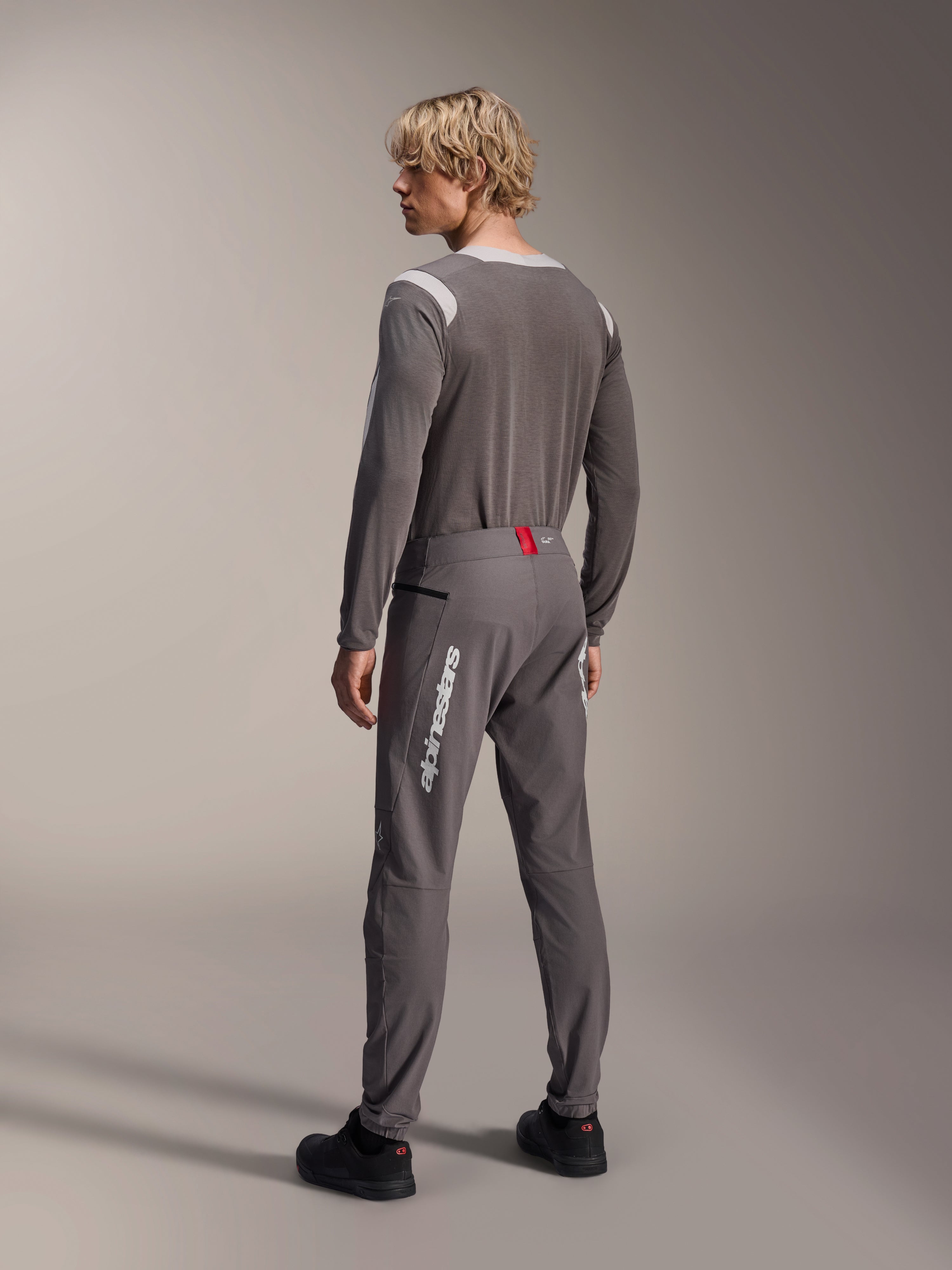 Model van achteren gezien in Alpinestars MTB-kleding, grijze A-Dura Dri Wool Jersey met lange mouwen, donkergrijze A-Dura Elite Pants met witte verticale branding en rood detail op de tailleband, gecoördineerde fietsoutfit voor mountainbiken