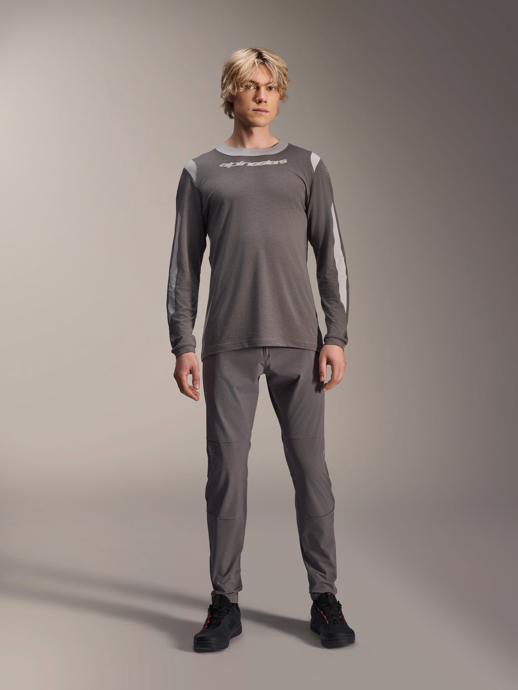 Alpinestars A-Dura Dri Wool Jersey met lange mouwen, donkergrijs, gedragen door model met bijpassende grijze broek en zwarte schoenen, witte Alpinestars-branding op de borst, lichtgrijze mesh-panelen op de schouders en mouwen, technische mountainbike-kleding