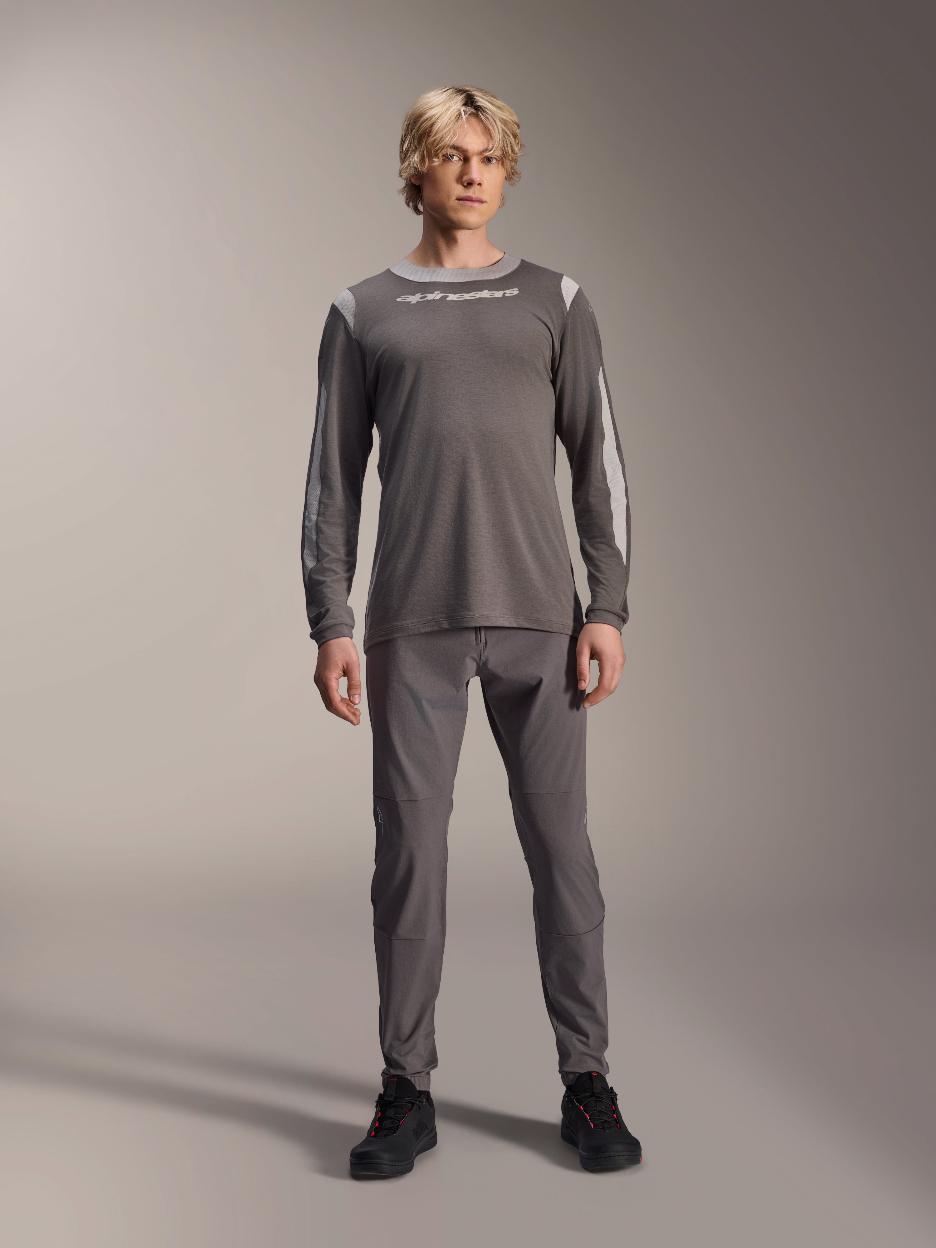 Alpinestars A-Dura Dri Wool Jersey met lange mouwen, donkergrijs, gedragen door model met bijpassende grijze broek en zwarte schoenen, witte Alpinestars-branding op de borst, lichtgrijze mesh-panelen op de schouders en mouwen, technische mountainbike-kleding