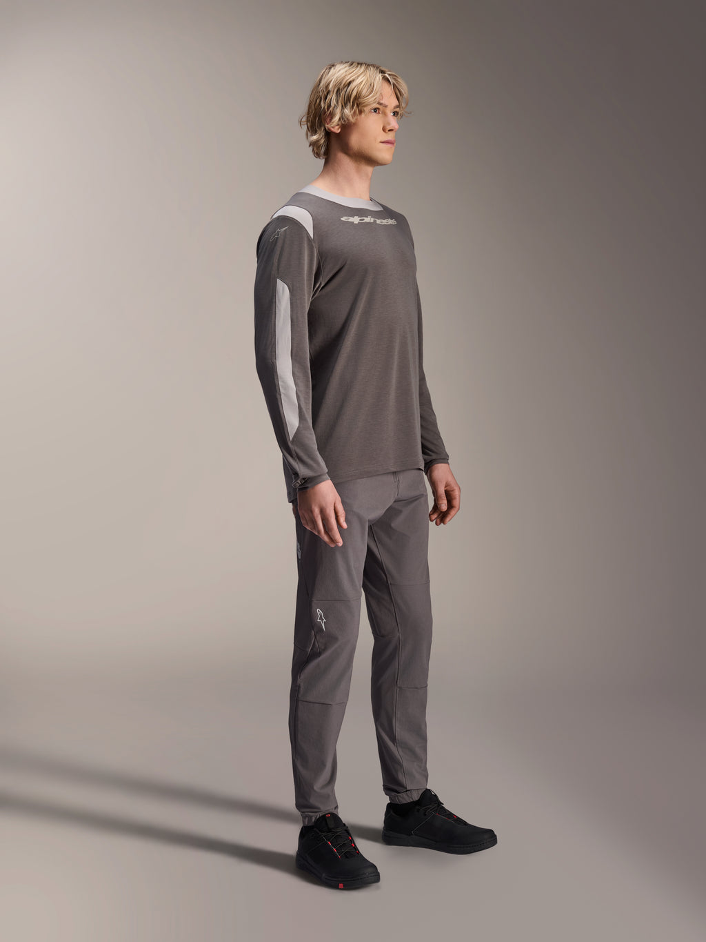 Mannelijk model draagt Alpinestars A-Dura Dri Wool Jersey met lange mouwen in donkergrijs, gecombineerd met bijpassende grijze broek en zwarte schoenen, voorzien van lichtgrijze mesh-panelen op de schouders en mouwen, technische MTB-uitrusting voor off-road fietsen