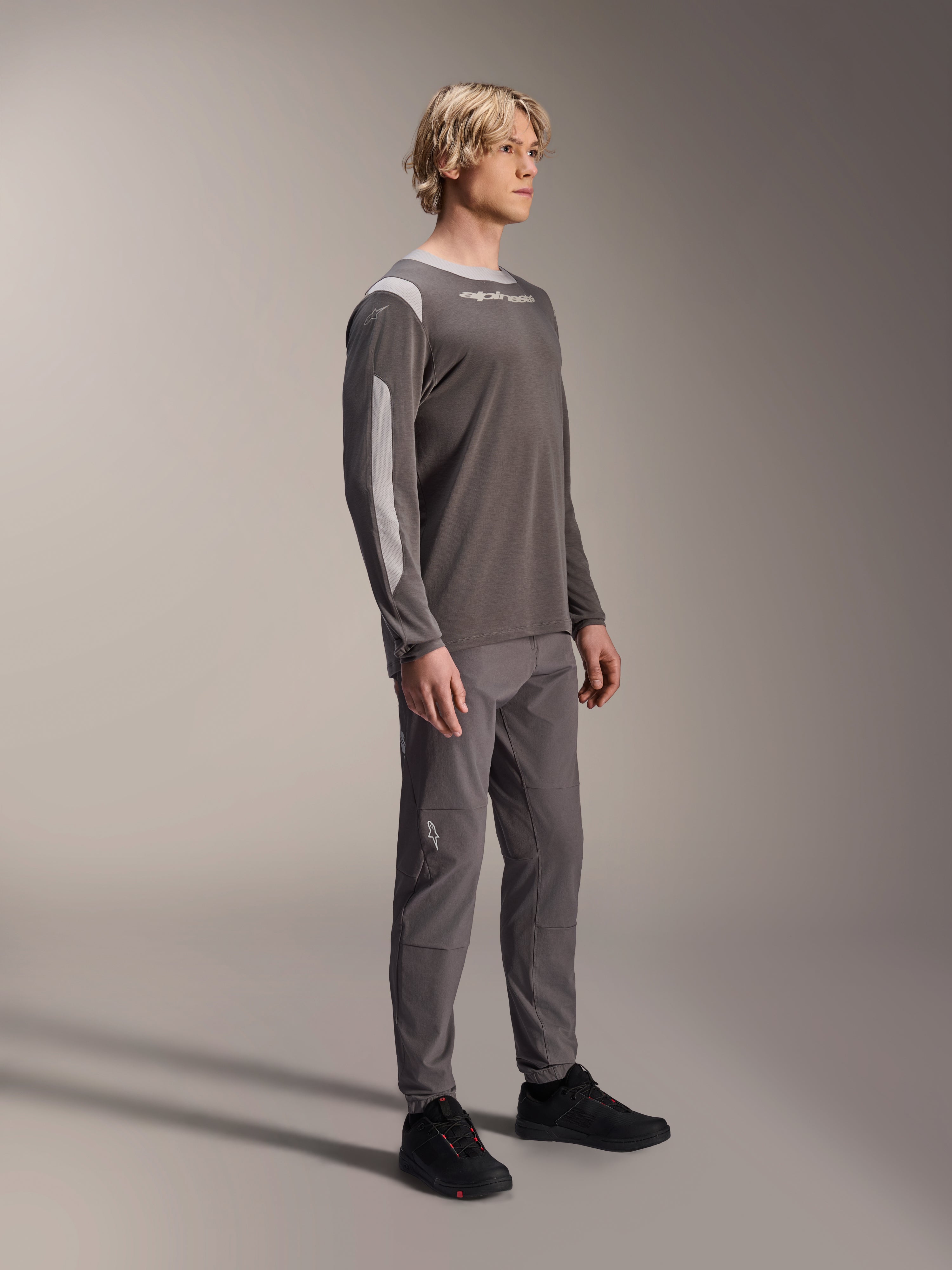 Mannelijk model draagt Alpinestars A-Dura Dri Wool Jersey met lange mouwen in donkergrijs, gecombineerd met bijpassende grijze broek en zwarte schoenen, voorzien van lichtgrijze mesh-panelen op de schouders en mouwen, technische MTB-uitrusting voor off-road fietsen