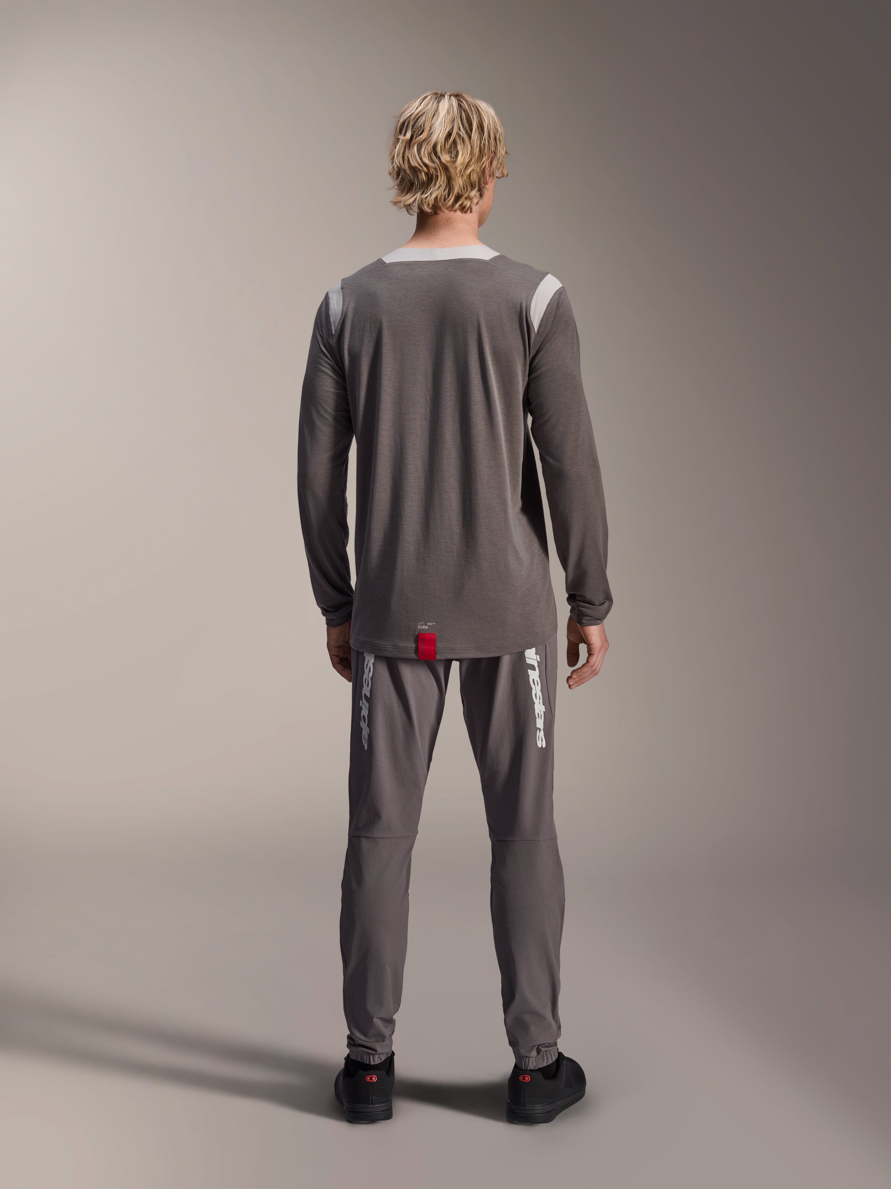 Alpinestars A-Dura Dri Wool Jersey - Lange Mouwen, donkergrijs, achteraanzicht op model, met lichtgrijze mesh-panelen op de schouders en kraag, verlengde achterzoom met een klein rood merklabel, gedragen met bijpassende grijze fietsbroek