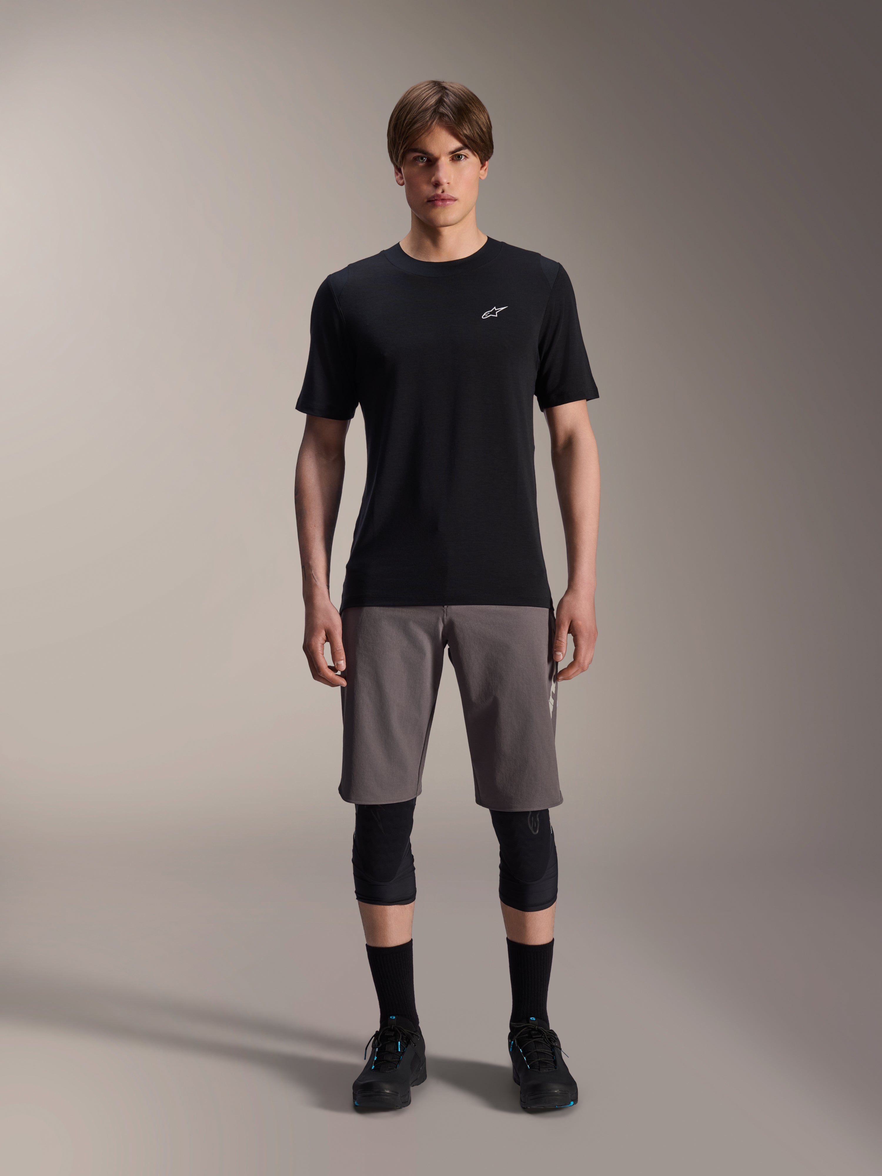 Alpinestars A-Dura Dri Wool Kurzarmtrikot, Schwarz, getragen von einem Model mit grauen Mountainbike-Shorts, schwarzen Knieprotektoren und schwarzen Fahrradschuhen, minimalistisches Design mit einem kleinen weißen Astars-Logo auf der Brust, funktionelle Performance-MTB-Bekleidung für Trail-Fahrten.