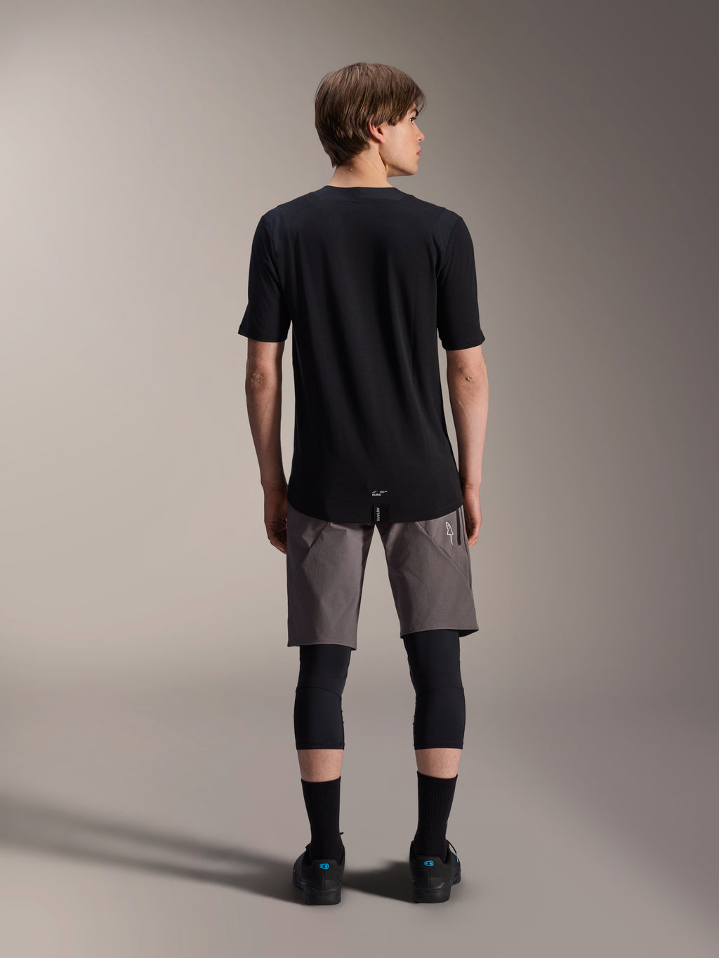 Alpinestars A-Dura Dri Wool Kurzarmtrikot, Schwarz, Rückansicht getragen von einem Modell mit grauen MTB-Shorts und schwarzen Knieschützern, mit längerem Rückensaum und athletischer Passform für den Offroad-Radsport