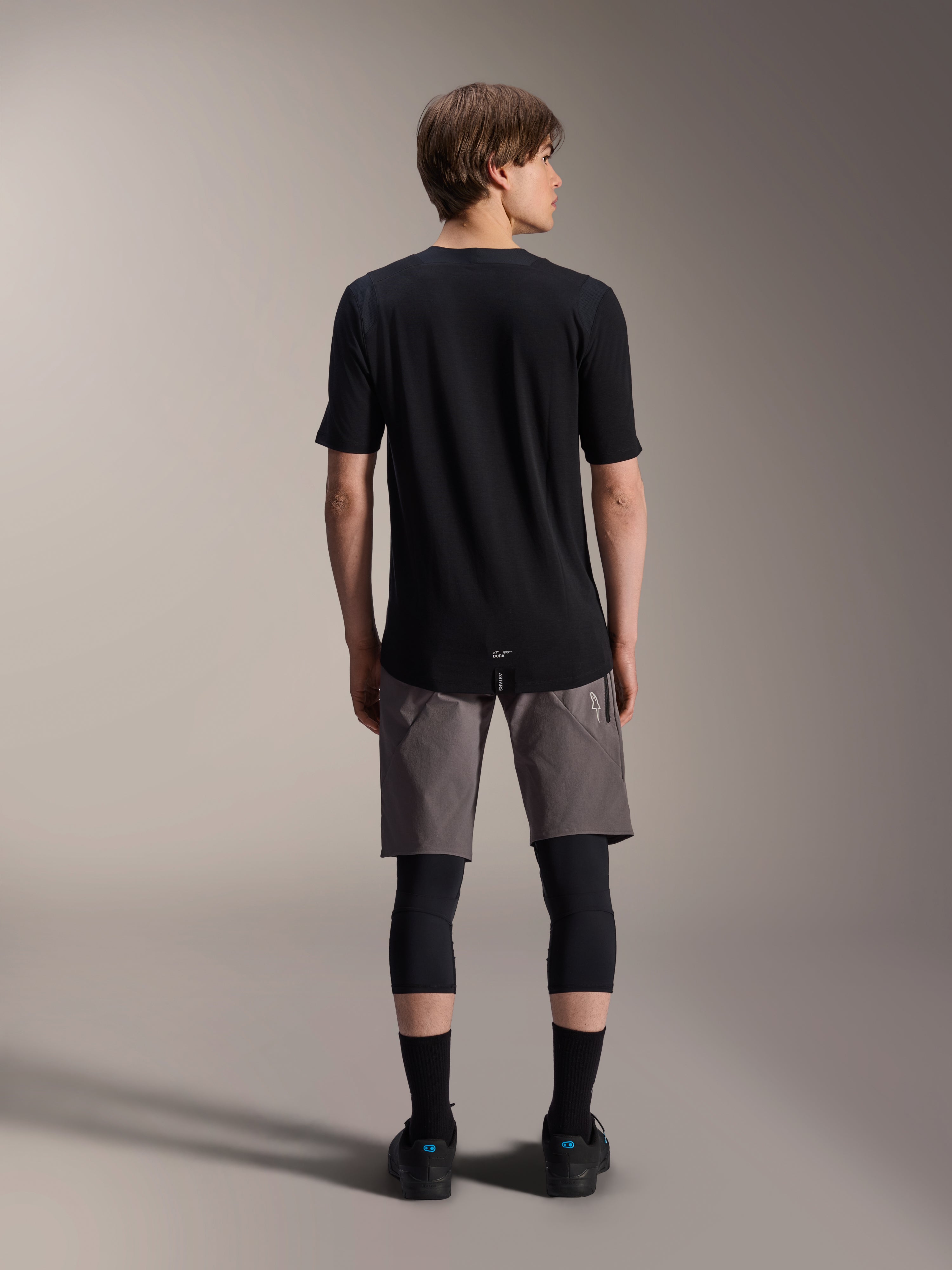 Alpinestars A-Dura Dri Wool Kurzarmtrikot, Schwarz, Rückansicht getragen von einem Modell mit grauen MTB-Shorts und schwarzen Knieschützern, mit längerem Rückensaum und athletischer Passform für den Offroad-Radsport