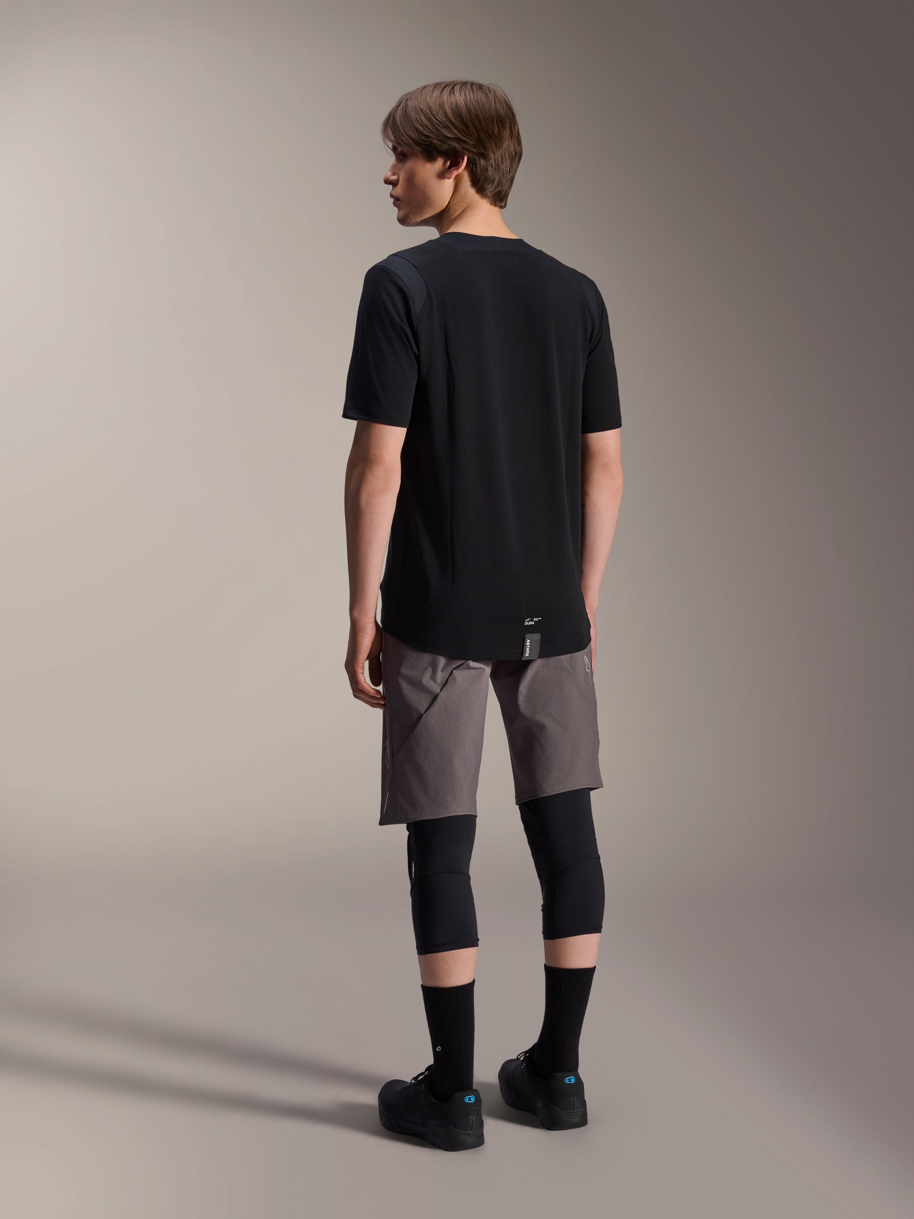 Alpinestars A-Dura Dri Wool Kurzarmtrikot und A-Dura Elite Shorts, schwarz- und dunkelgraue Mountainbike-Bekleidung, getragen mit schwarzen Knieschützern und Socken, Rückansicht der Radsportausrüstung für Offroad-Performance