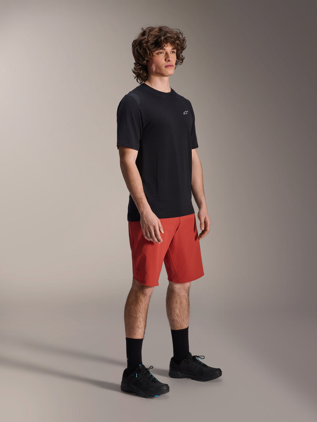 Männliches Model in Alpinestars A-Dura Elite Shorts in Dark Orange und A-Dura Dri Wool Kurzarm-Trikot in Schwarz, Mountainbike-Performance-Ausrüstung, kombiniert mit schwarzen Socken und Schuhen, sportliches Design für Offroad-Radsport