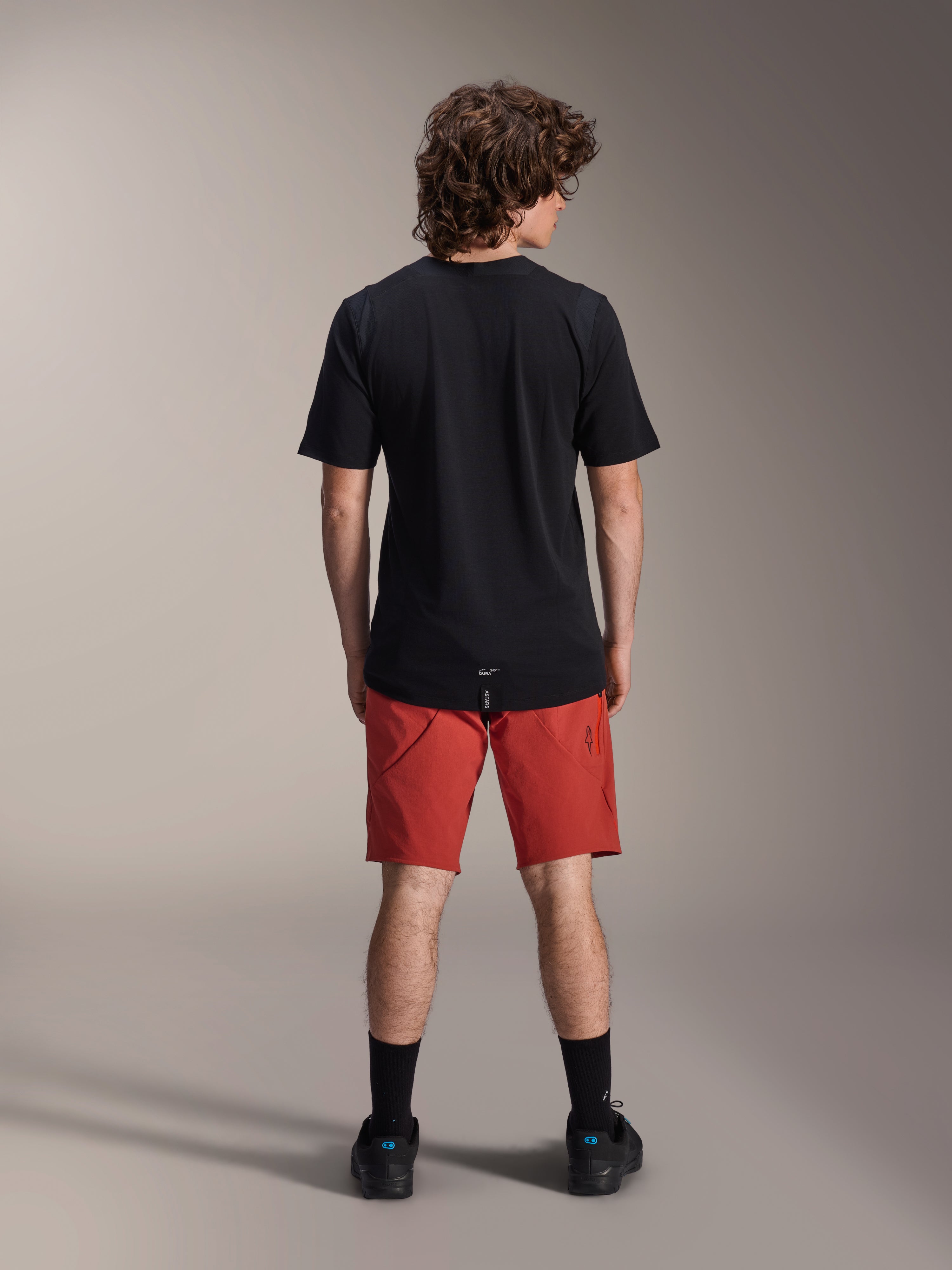 Männliches Model in Alpinestars A-Dura Elite Shorts in Dark Orange und schwarzem A-Dura Dri Wool Kurzarmtrikot, Rückansicht, kombiniert mit schwarzen Socken und Fahrradschuhen, professionelle Mountainbike-Bekleidung