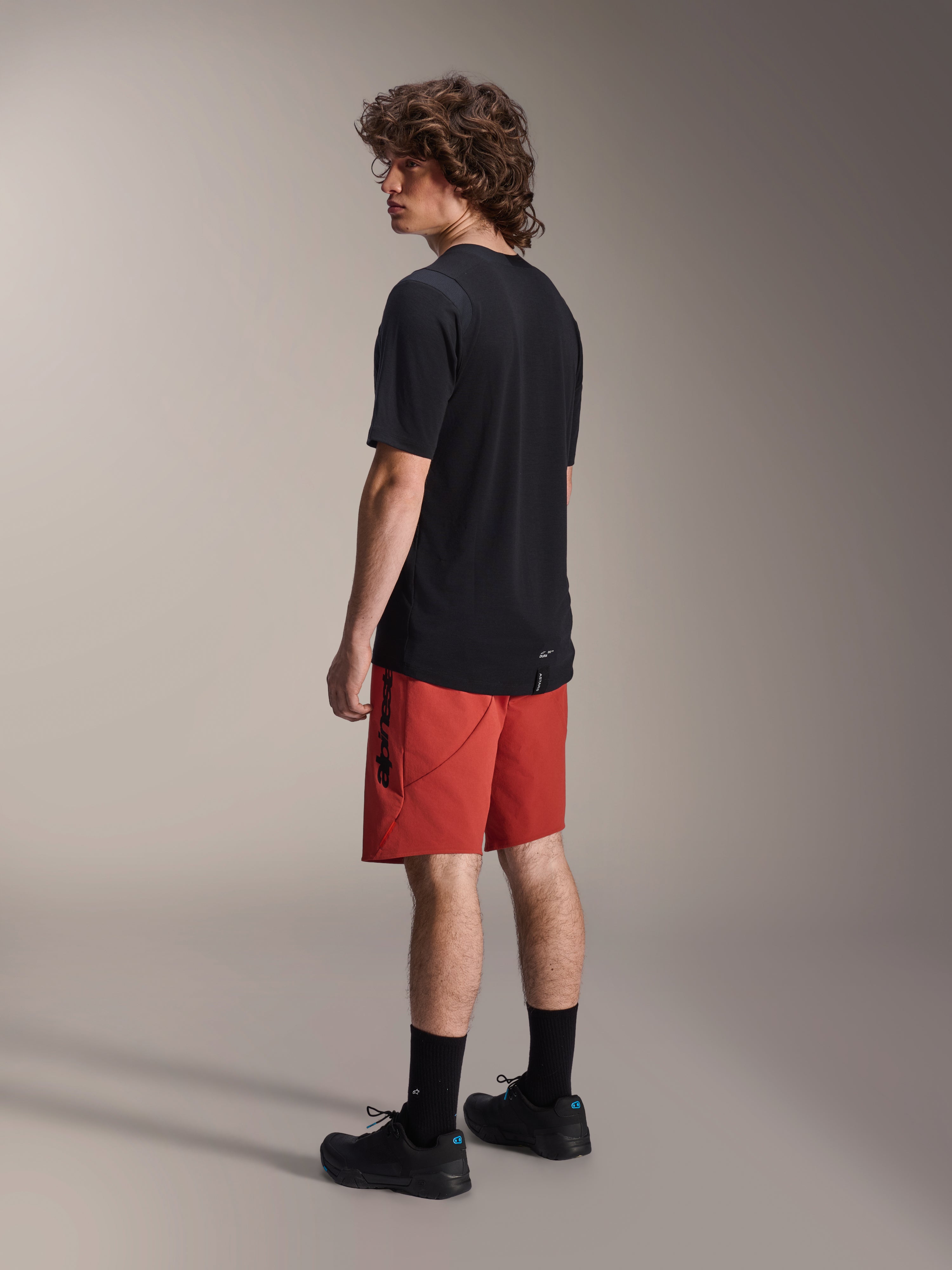Alpinestars A-Dura Elite Shorts, Mountainbike-Shorts, Dunkelorange, kombiniert mit einem schwarzen kurzärmeligen Trikot und schwarzen Mountainbike-Schuhen, rückseitige Seitenansicht des Modells zur Darstellung der Performance-Passform und des Brandings