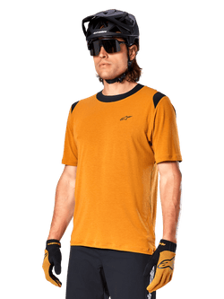 Maillot Alpinestars A-Dura Dri Wool à manches courtes, maillot de cyclisme VTT, Or foncé avec accents noirs, maillot technique à manches courtes en mélange de laine avec empiècements noirs aux épaules, porté avec un casque de VTT noir et des gants assortis