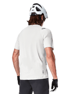 Alpinestars A-Aria Astar Jersey, manches courtes, White Dove, vue arrière d'un maillot de VTT léger et respirant, porté avec un casque blanc et des gants de cyclisme noirs, doté d'un pan arrière plus long pour une couverture accrue