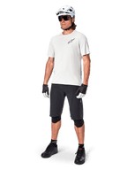 Cycliste VTT portant un maillot à manches courtes Alpinestars A-Aria Astar en White Dove, associé à un short de VTT noir, un casque blanc et des genouillères noires, avec un petit logo noir sur la poitrine, équipement de cyclisme coordonné pour la performance sur sentier