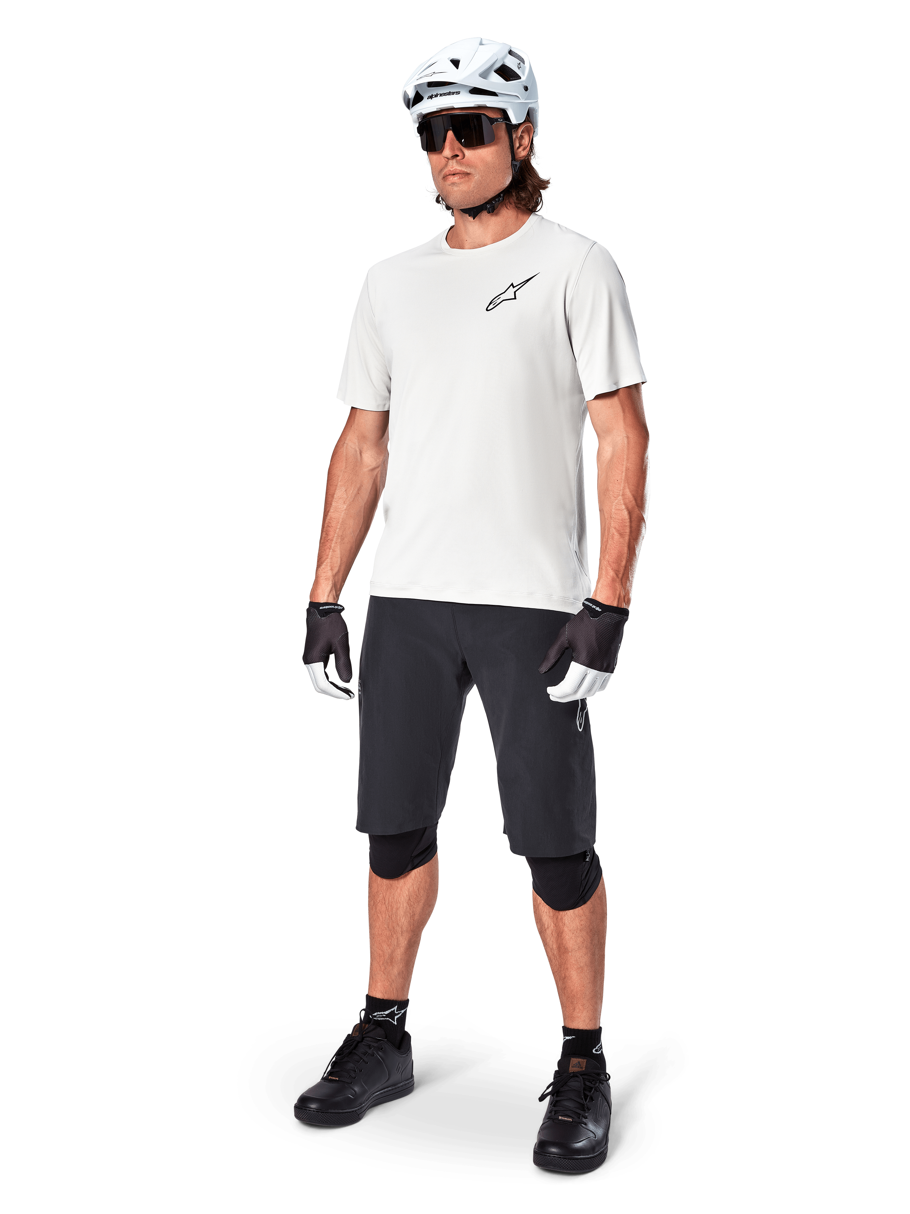 Cycliste VTT portant un maillot à manches courtes Alpinestars A-Aria Astar en White Dove, associé à un short de VTT noir, un casque blanc et des genouillères noires, avec un petit logo noir sur la poitrine, équipement de cyclisme coordonné pour la performance sur sentier