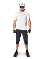 Cycliste portant le maillot à manches courtes Alpinestars A-Aria Astar en coloris White Dove, avec un logo Astars noir sur la poitrine, assorti d'un short de VTT noir, d'un casque blanc, de lunettes de soleil, de genouillères noires et de gants, tenue de cyclisme tout-terrain complète.