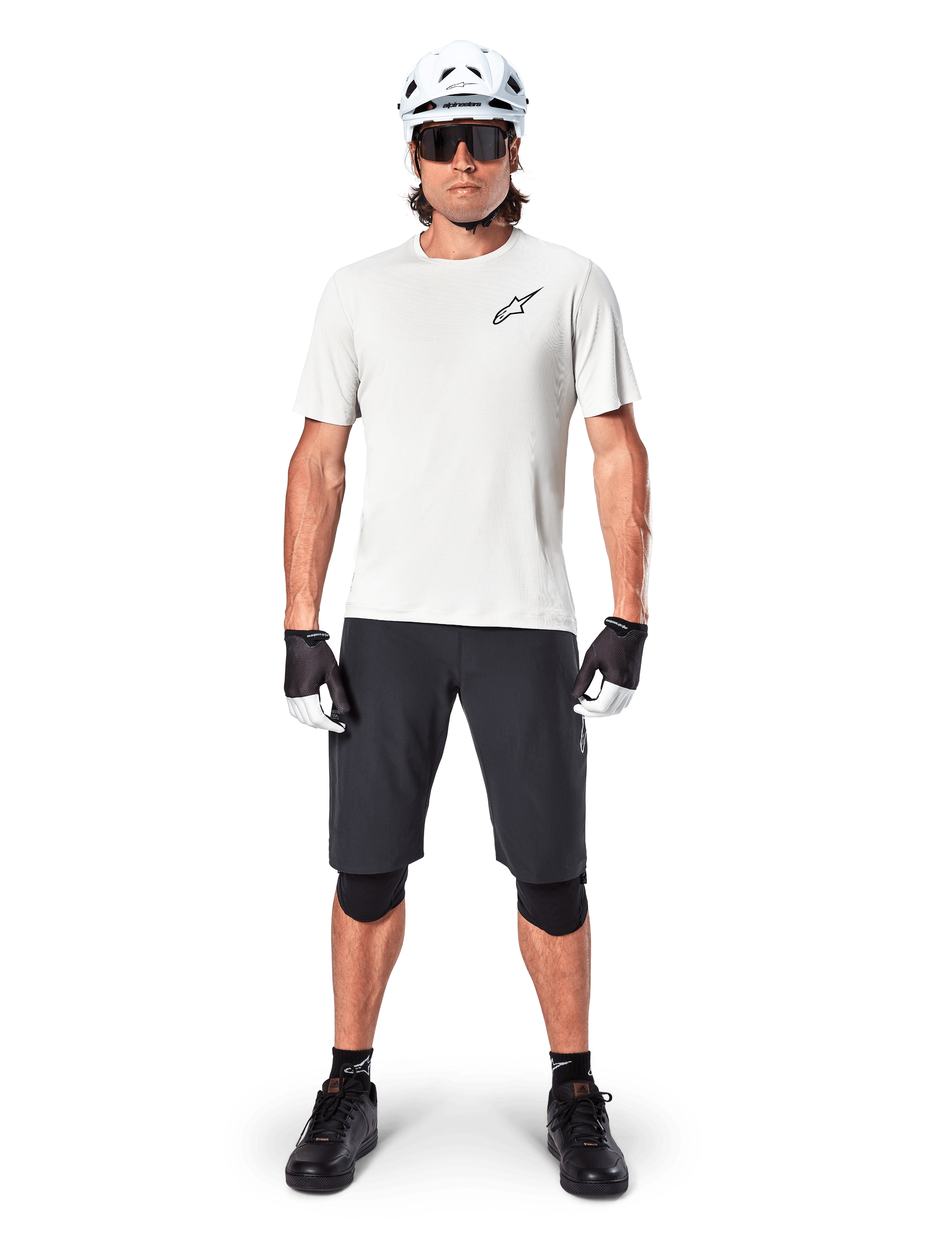 Cycliste portant le maillot à manches courtes Alpinestars A-Aria Astar en coloris White Dove, avec un logo Astars noir sur la poitrine, assorti d'un short de VTT noir, d'un casque blanc, de lunettes de soleil, de genouillères noires et de gants, tenue de cyclisme tout-terrain complète.