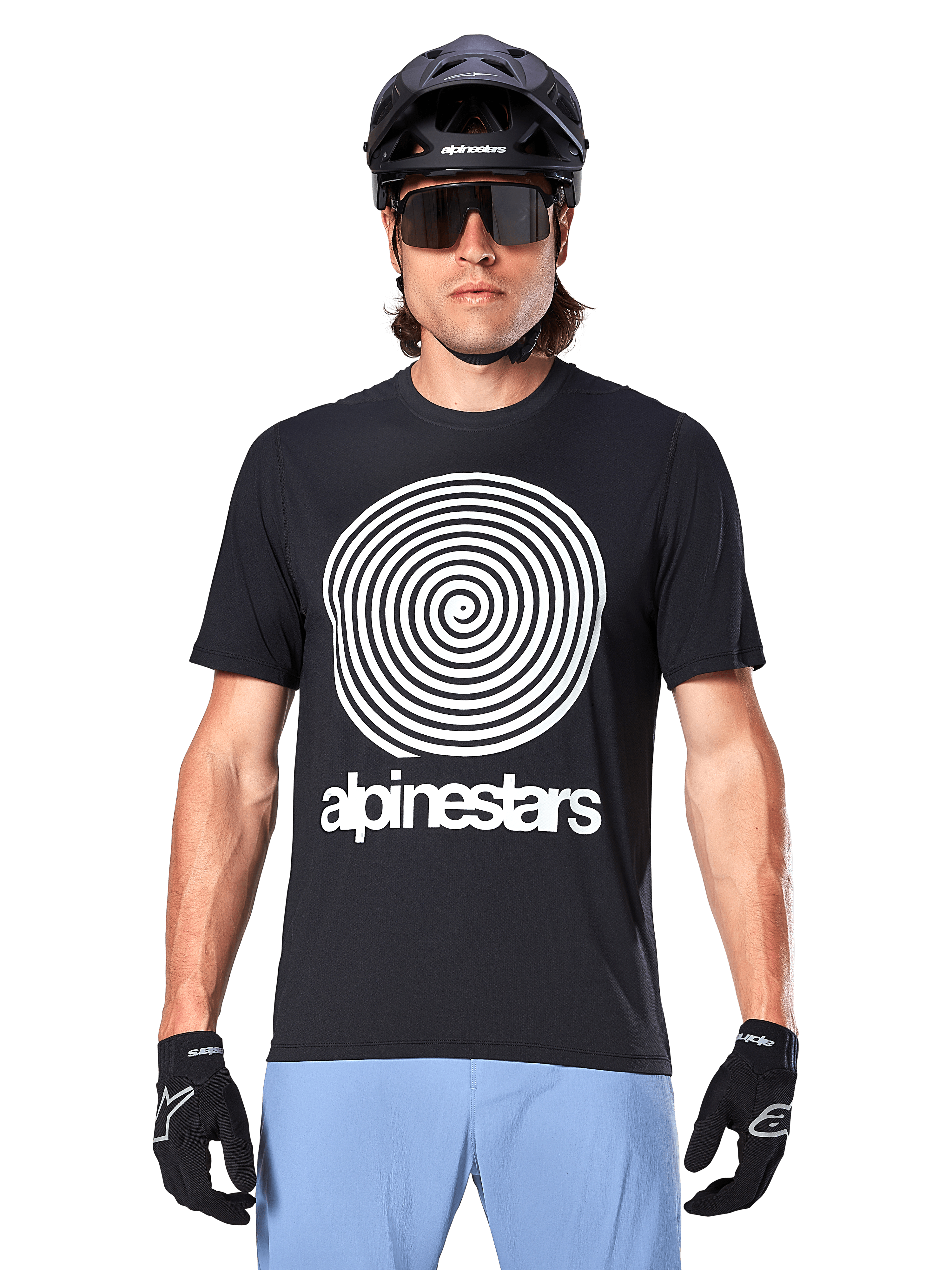 Maillot Alpinestars A-Dura Oscar, maillot de VTT à manches courtes, noir, avec un large graphique en spirale Oscar vintage blanc sur la poitrine, porté avec un casque de vélo noir, des lunettes de soleil, des gants noirs et un short bleu clair, équipement de cyclisme tout-terrain d'inspiration héritage