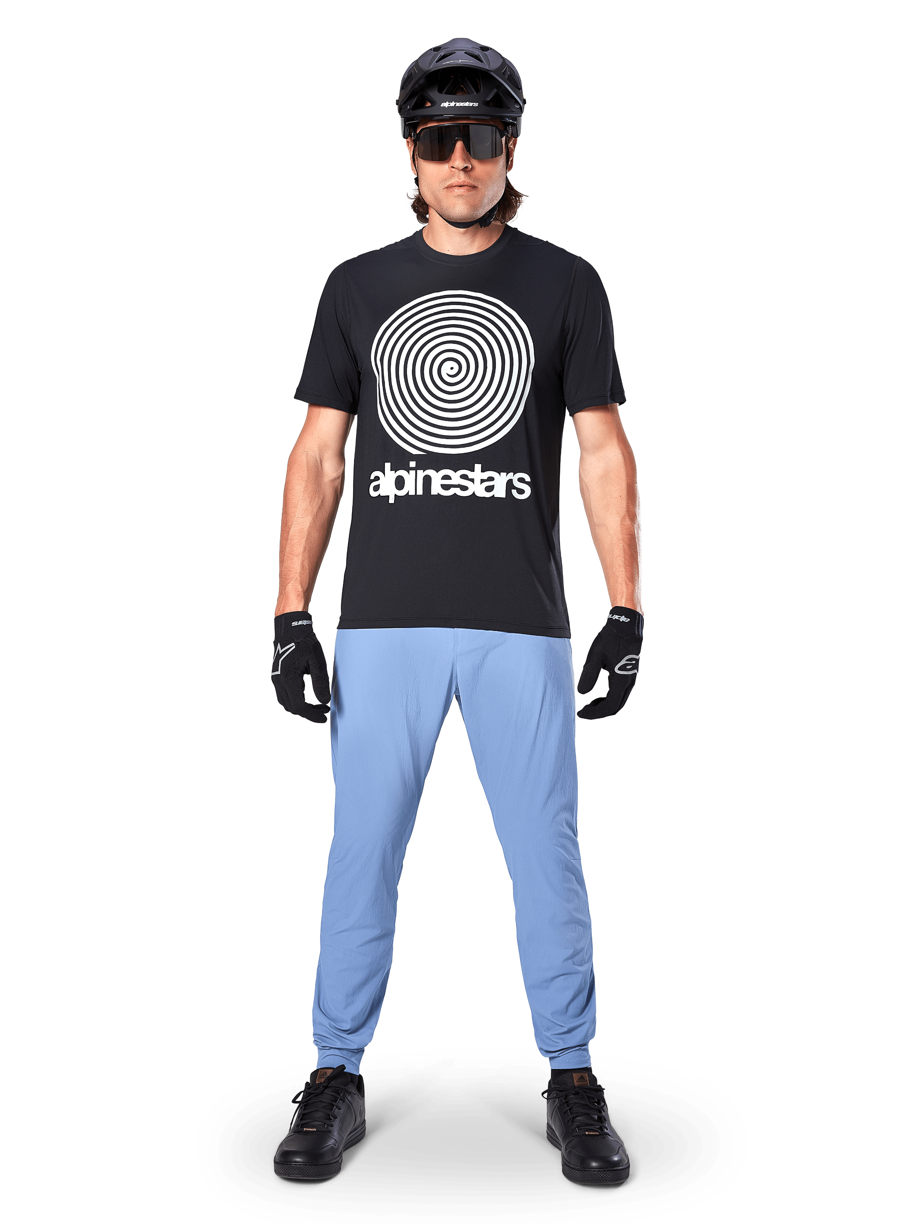 Maillot à manches courtes Alpinestars A-Dura Oscar, maillot de VTT noir avec un grand graphique en spirale Oscar vintage blanc et le nom de la marque, porté avec un pantalon bleu clair, un casque noir, des lunettes de soleil et des gants noirs, équipement de performance pour le VTT