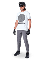 Maillot à manches courtes Alpinestars A-Dura Oscar, blanc White Dove avec grand graphique en spirale noire, porté par un cycliste avec pantalon de VTT gris, casque noir et gants, équipement de cyclisme de performance pour le tout-terrain