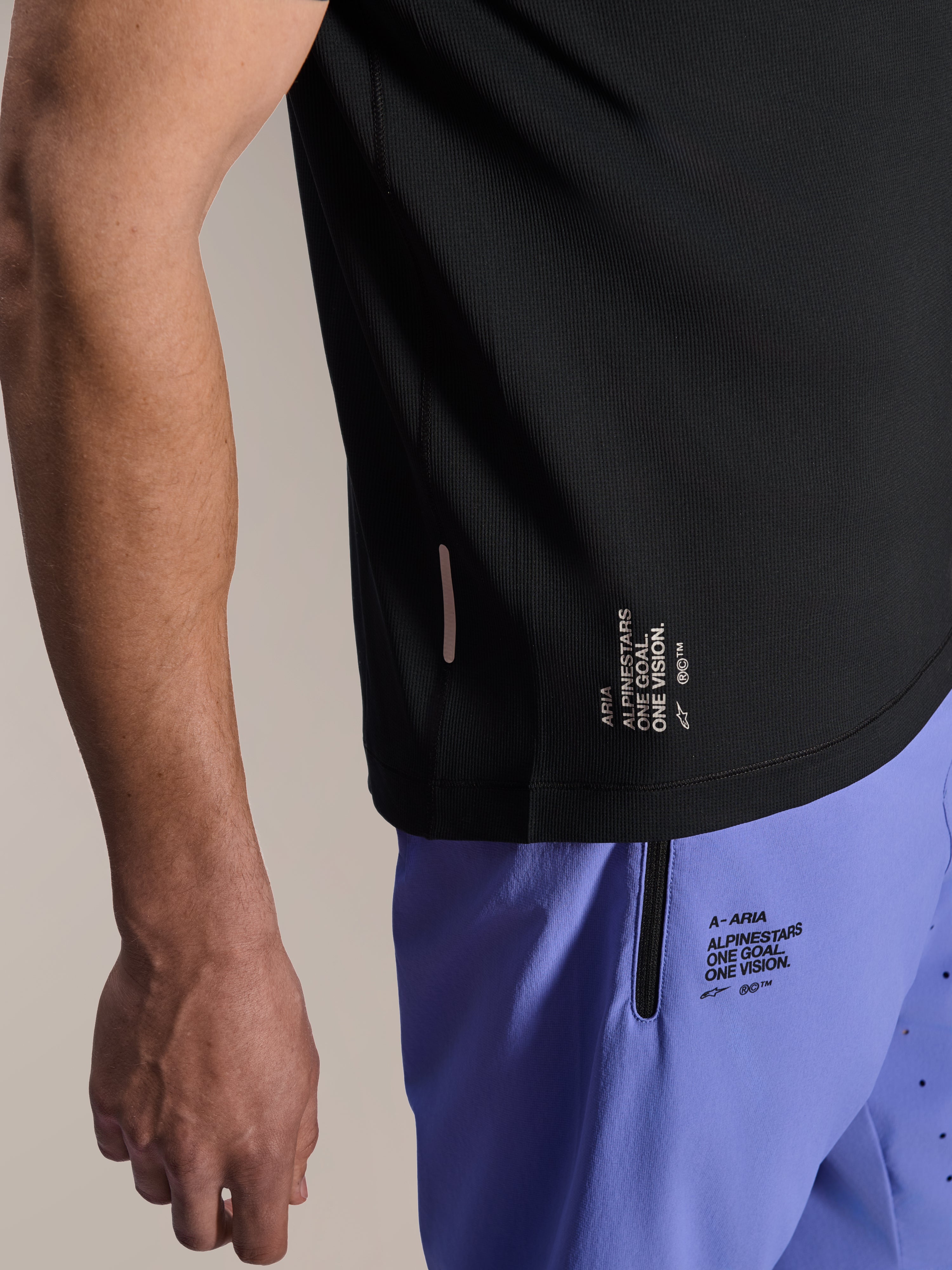 Maillot à manches courtes Alpinestars A-Aria Switch, maillot de VTT noir, gros plan sur l'ourlet latéral avec le marquage blanc « ONE GOAL. ONE VISION. », porté avec un short de cyclisme violet clair doté de logos assortis.