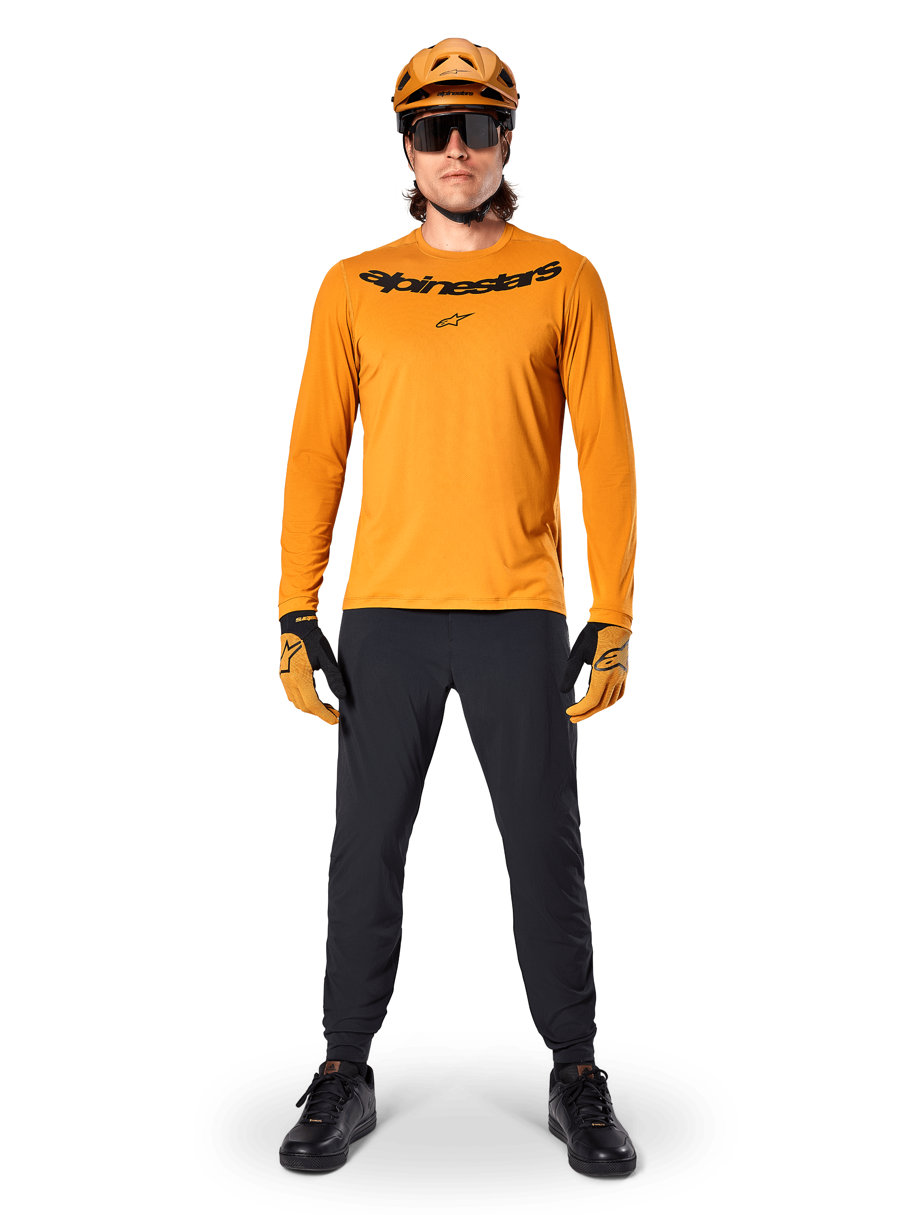 Alpinestars A-Dura Rocker Langarmtrikot in Dark Gold, mit schwarzem Siebdruck-Logo auf der Brust, getragen mit einem passenden goldfarbenen MTB-Helm, schwarzer Sonnenbrille und schwarzer Hose, professionelle Mountainbike-Ausrüstung