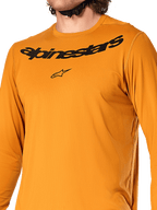 Alpinestars A-Dura Rocker Langarmtrikot, Dunkelgold, Nahaufnahme des Brustbereichs mit schwarzem Alpinestars-Schriftzug und Stern-Logo, strukturiertes feuchtigkeitsableitendes Jacquard-Gewebe, Performance-MTB-Radsportbekleidung