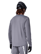 Alpinestars A-Dura Rocker Jersey met lange mouwen in donkergrijs, achteraanzicht van een mountainbiker, gecombineerd met een bijpassende grijze short met witte branding, zwarte MTB-helm en zwarte handschoenen, professionele off-road fietsuitrusting