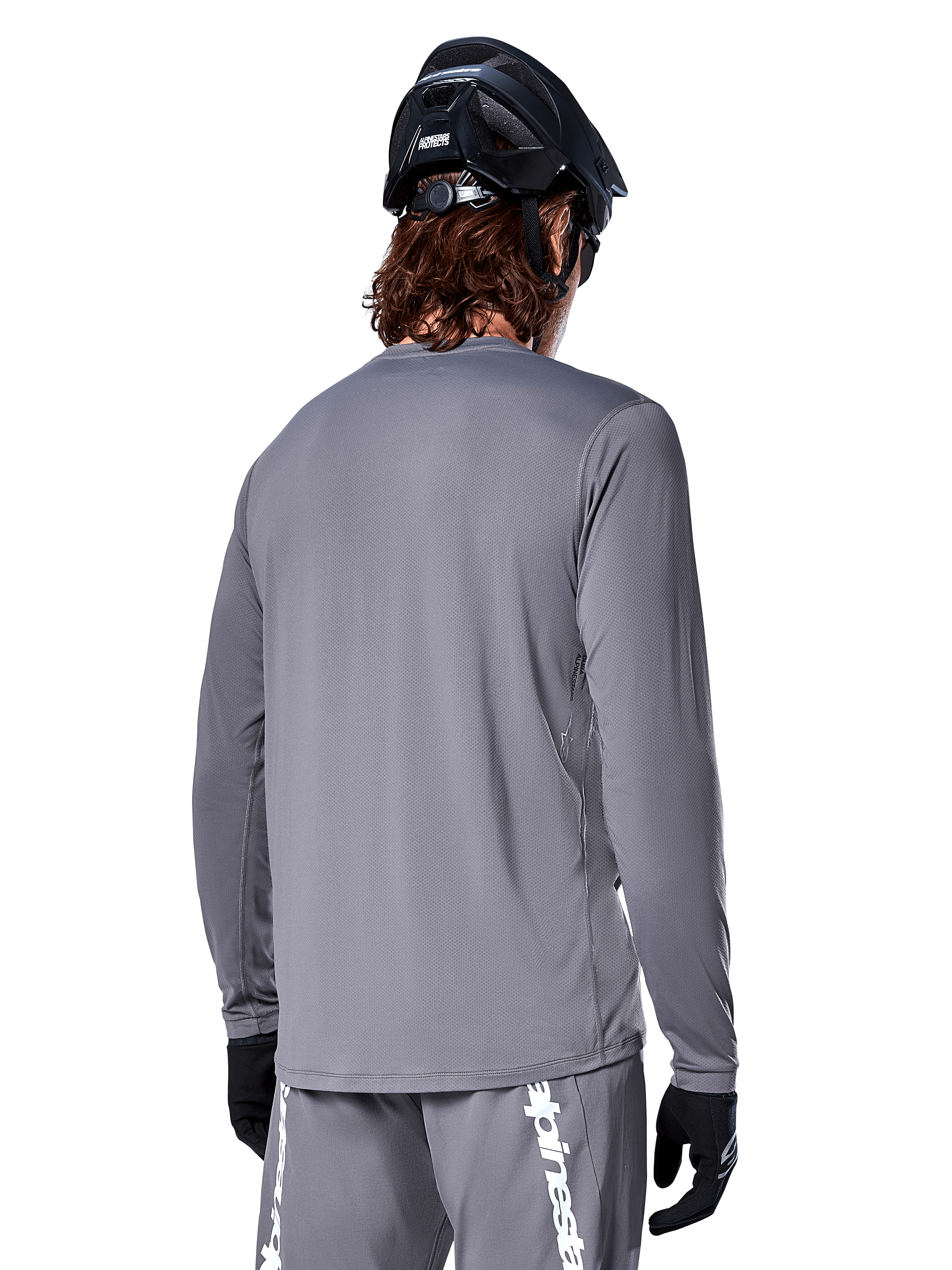 Alpinestars A-Dura Rocker Jersey met lange mouwen in donkergrijs, achteraanzicht van een mountainbiker, gecombineerd met een bijpassende grijze short met witte branding, zwarte MTB-helm en zwarte handschoenen, professionele off-road fietsuitrusting