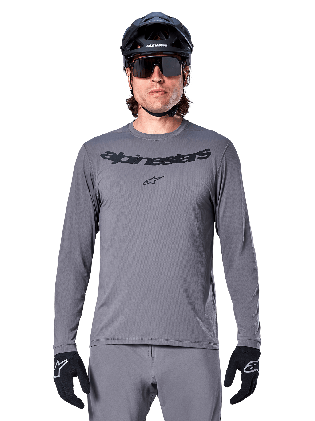 Mannelijke rijder in het Alpinestars A-Dura Rocker Jersey met lange mouwen, donkergrijze MTB-jersey met zwart woordmerk op de borst, gecombineerd met grijze broek, zwarte helm en handschoenen, ontworpen voor off-road fietsen