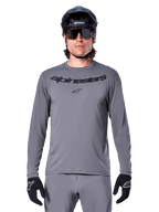 Mannelijke rijder in het Alpinestars A-Dura Rocker Jersey met lange mouwen, donkergrijze MTB-jersey met zwart woordmerk op de borst, gecombineerd met grijze broek, zwarte helm en handschoenen, ontworpen voor off-road fietsen
