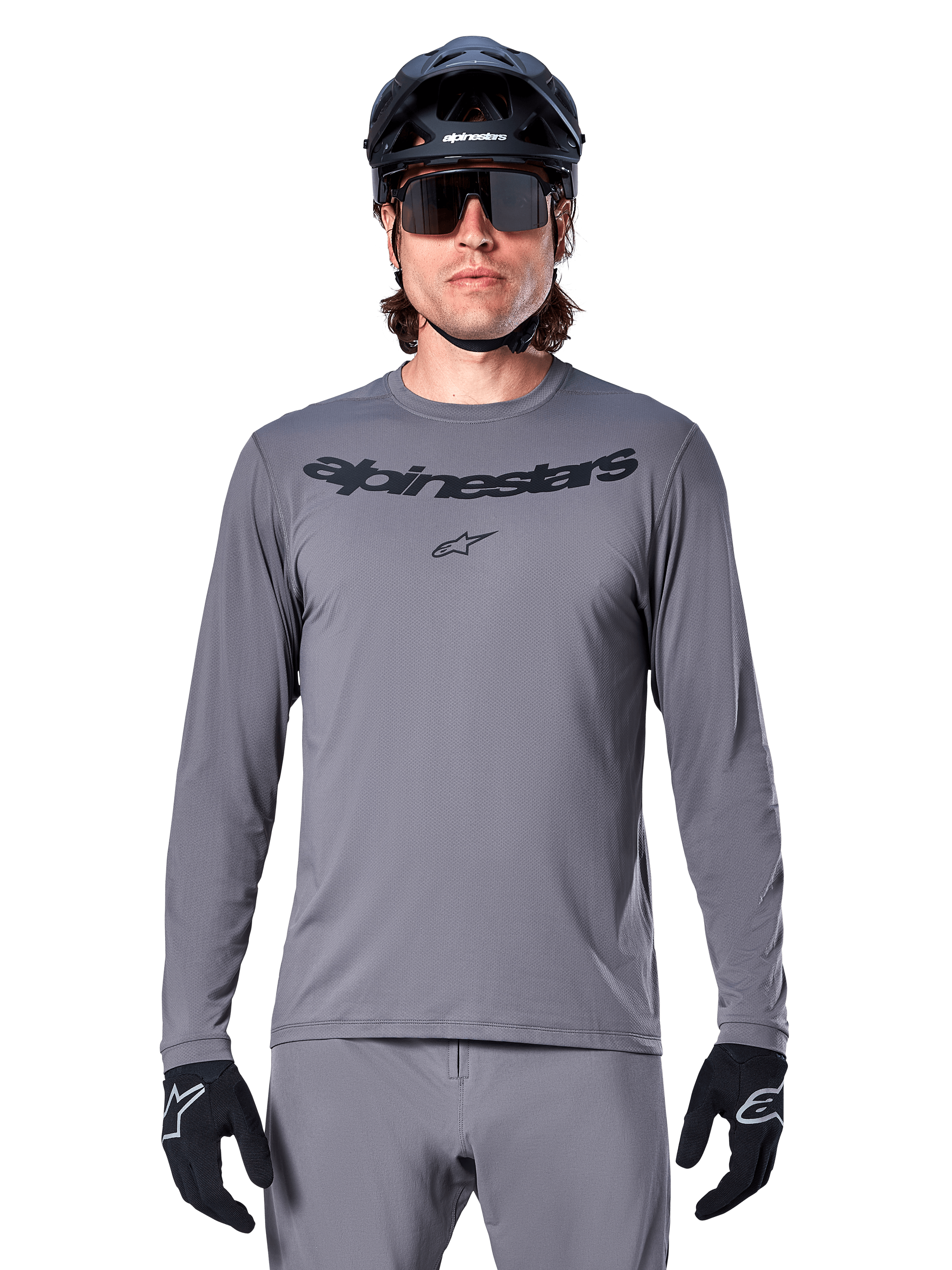 Mannelijke rijder in het Alpinestars A-Dura Rocker Jersey met lange mouwen, donkergrijze MTB-jersey met zwart woordmerk op de borst, gecombineerd met grijze broek, zwarte helm en handschoenen, ontworpen voor off-road fietsen