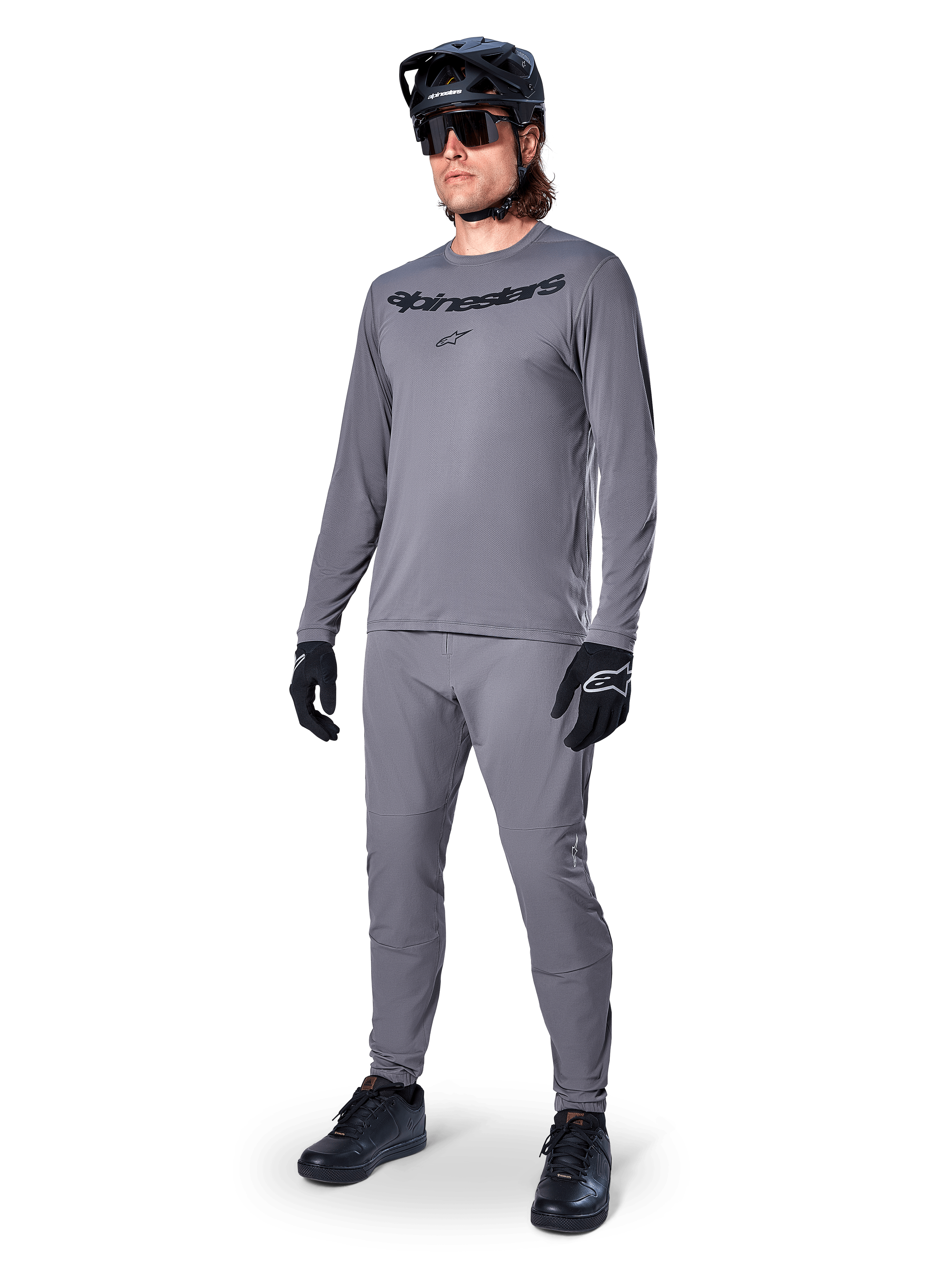 Alpinestars A-Dura Rocker Jersey met lange mouwen, donkergrijs, gedragen door een model met bijpassende grijze broek, zwarte mountainbikehelm, zonnebril en handschoenen, casual MTB-stijl voor off-road rijden