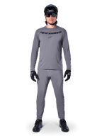 Mannelijke fietser in Alpinestars A-Dura Rocker Jersey met lange mouwen in donkergrijs, bijpassende grijze broek, zwarte MTB-helm, zonnebril en handschoenen, professionele mountainbikekleding voor off-road paden